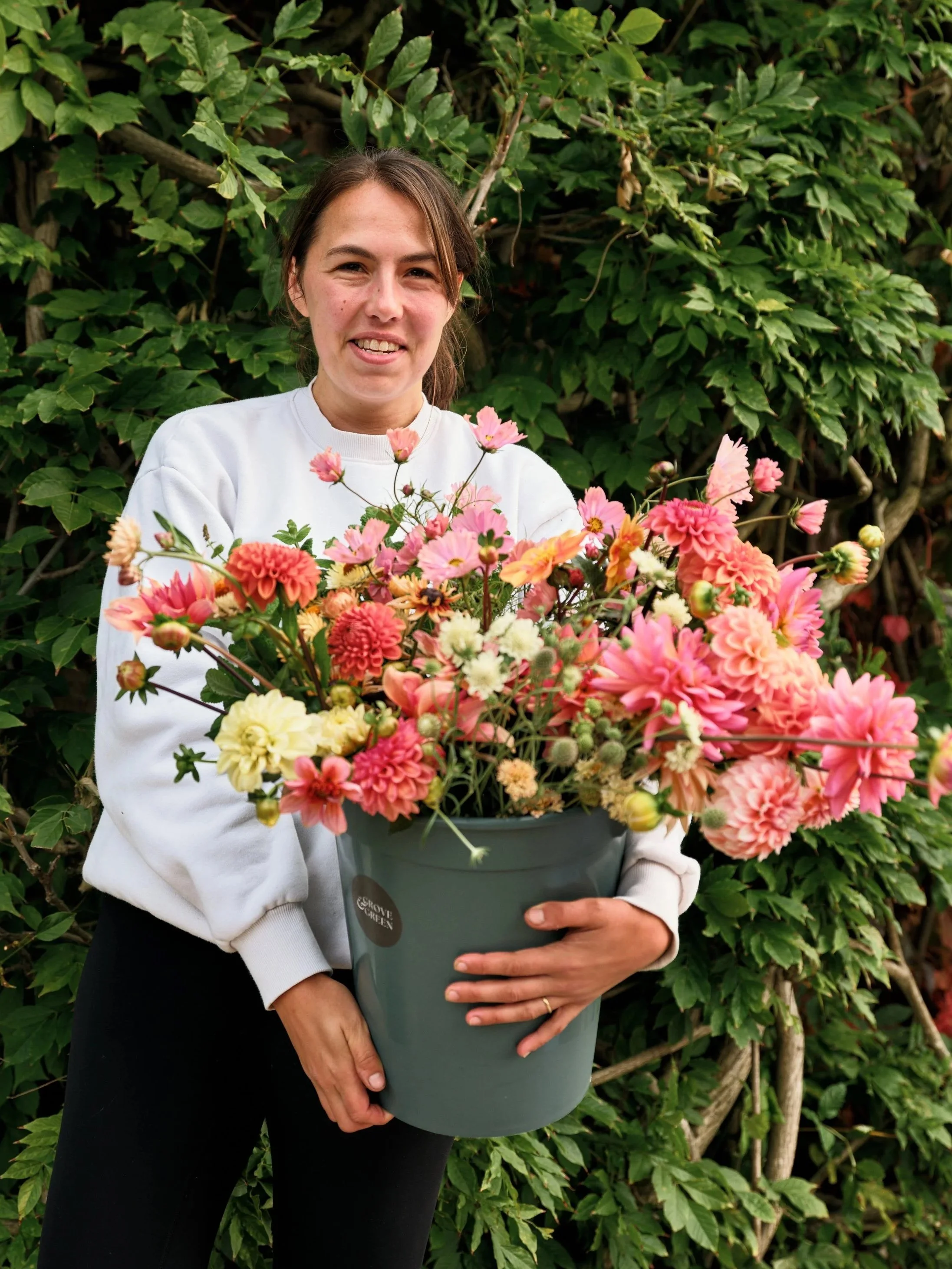 A woman in a white sweatshirt holding a large gray pot filled with pink, white, and peach flowers, standing in front of lush green foliage.
