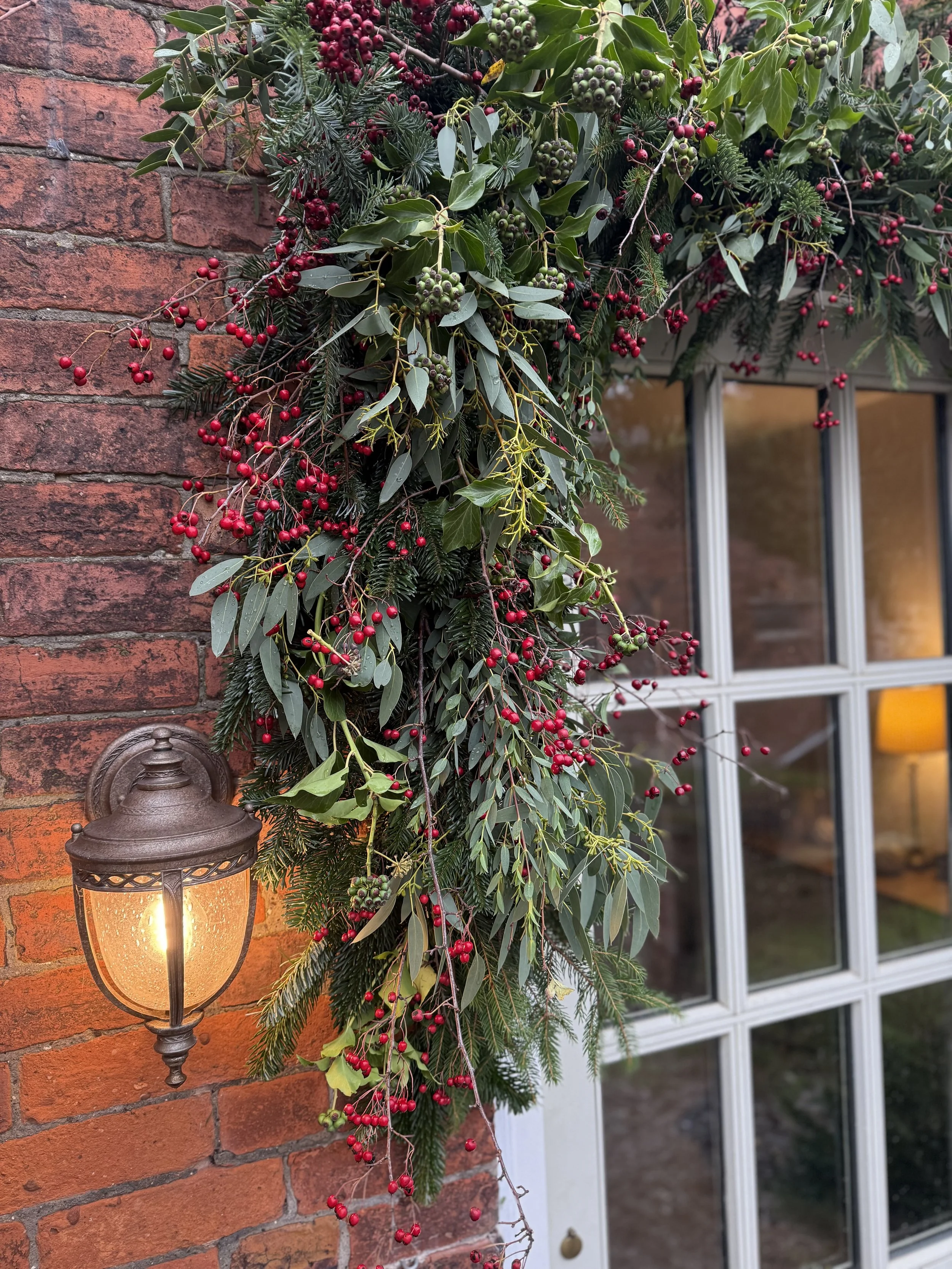 A holiday decoration arrangement of evergreen branches, holly leaves, and red berries hanging on a brick wall beside a window, illuminated by a vintage outdoor lantern.