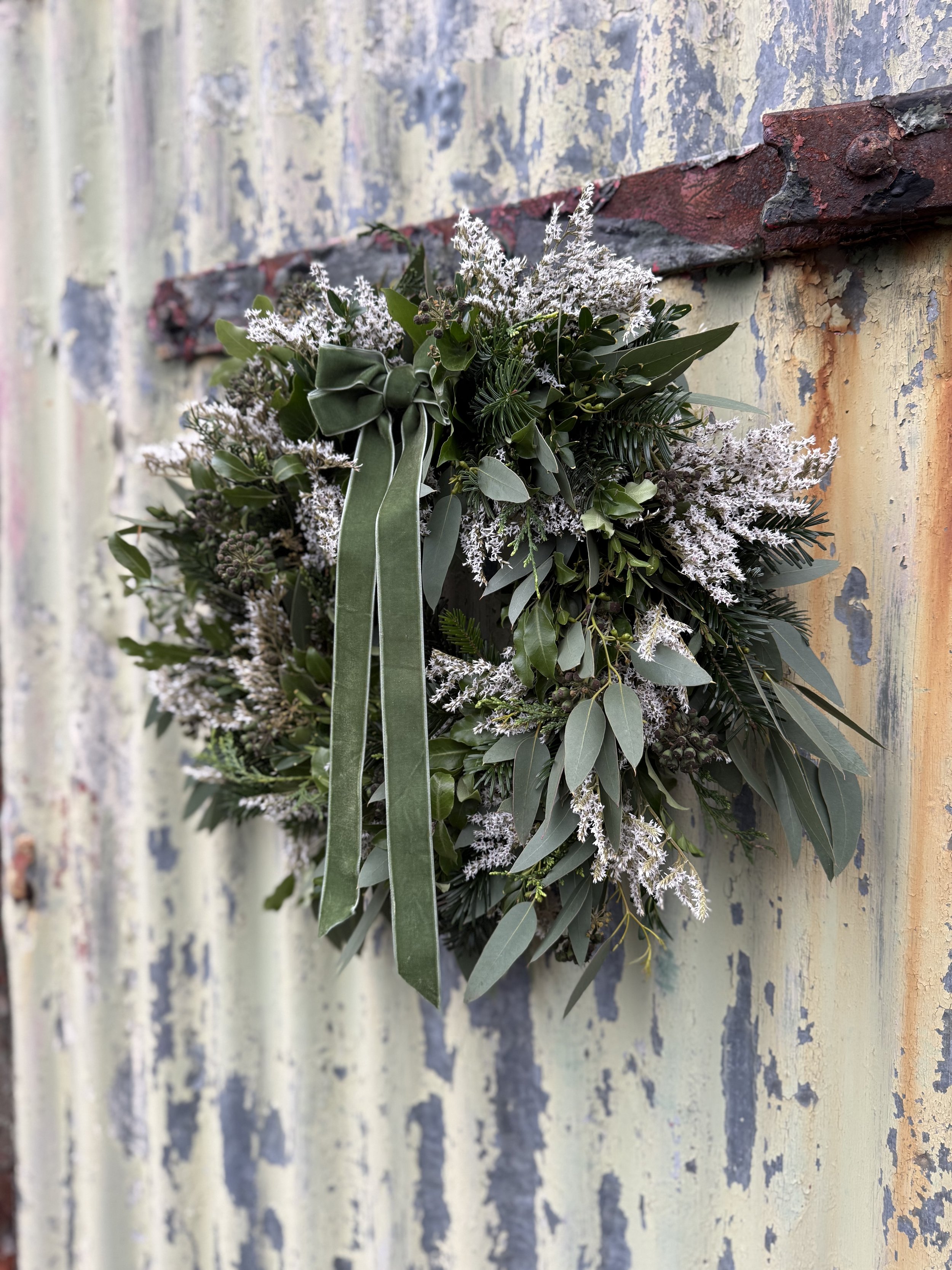 A decorative wreath made of green foliage, white flowers, and a large green bow, hanging on a weathered, chipped, painted metal surface.