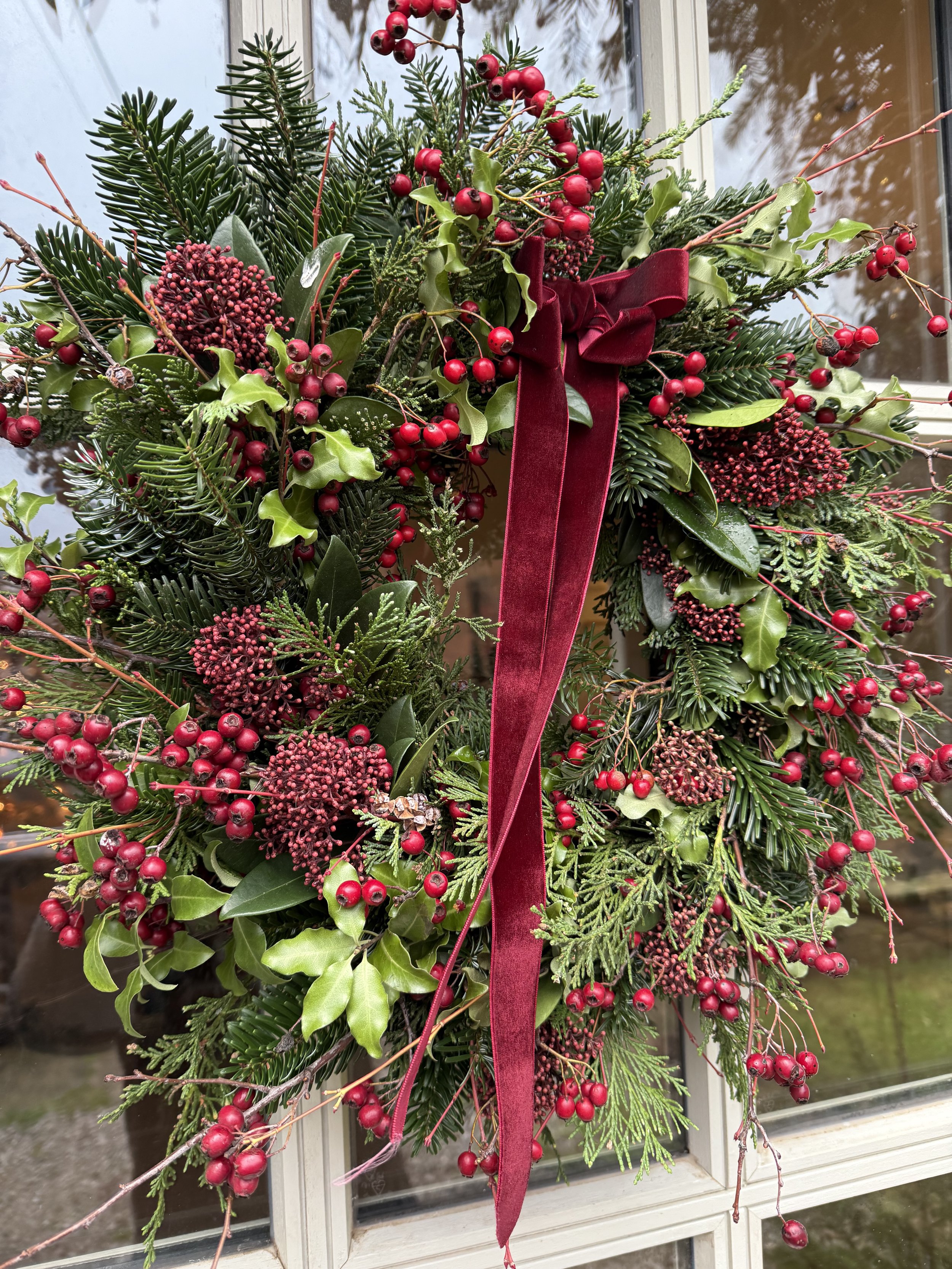 A Christmas wreath decorated with red berries, green holly leaves, and pine branches, adorned with a large red velvet ribbon, hanging on a window.