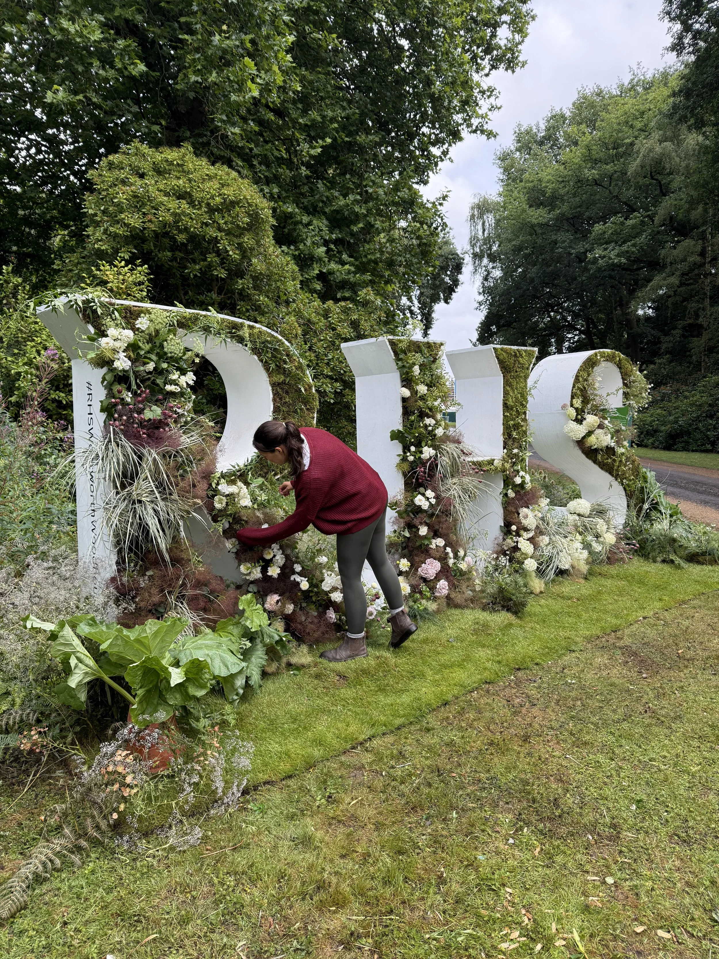 A woman arranging flowers in front of large white letters spelling 'LOVE' decorated with flowers and greenery, outdoors in a lush green park.