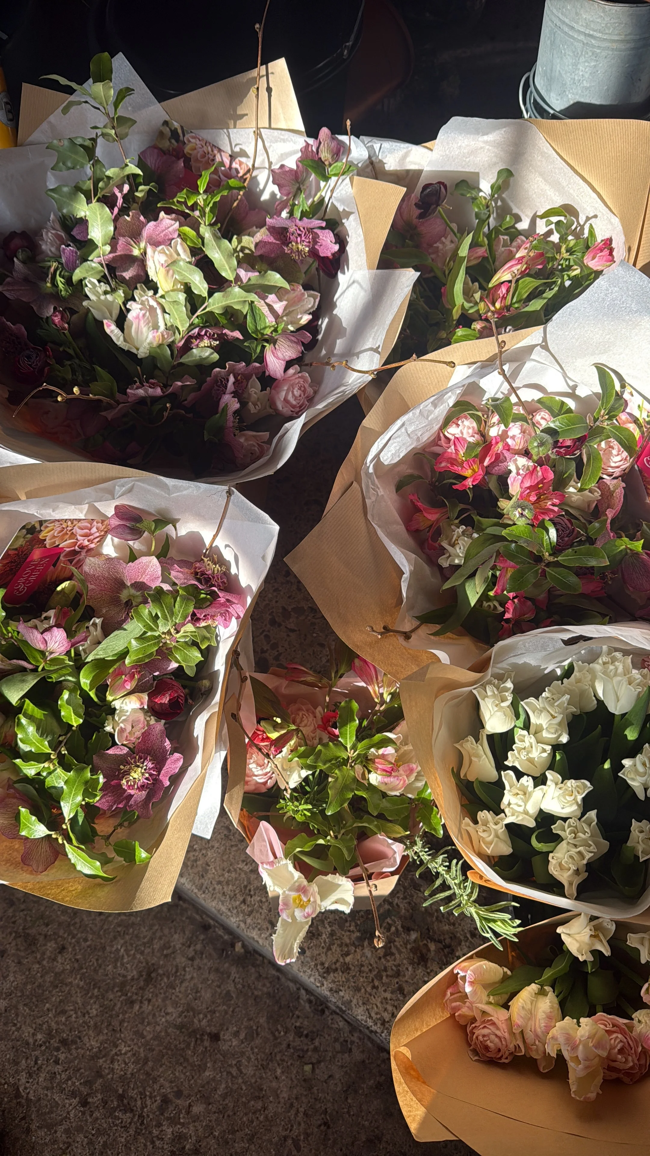 Multiple bouquets of mixed pink, white, and purple flowers with green leaves, wrapped in white and brown paper ready for Mother's Day.