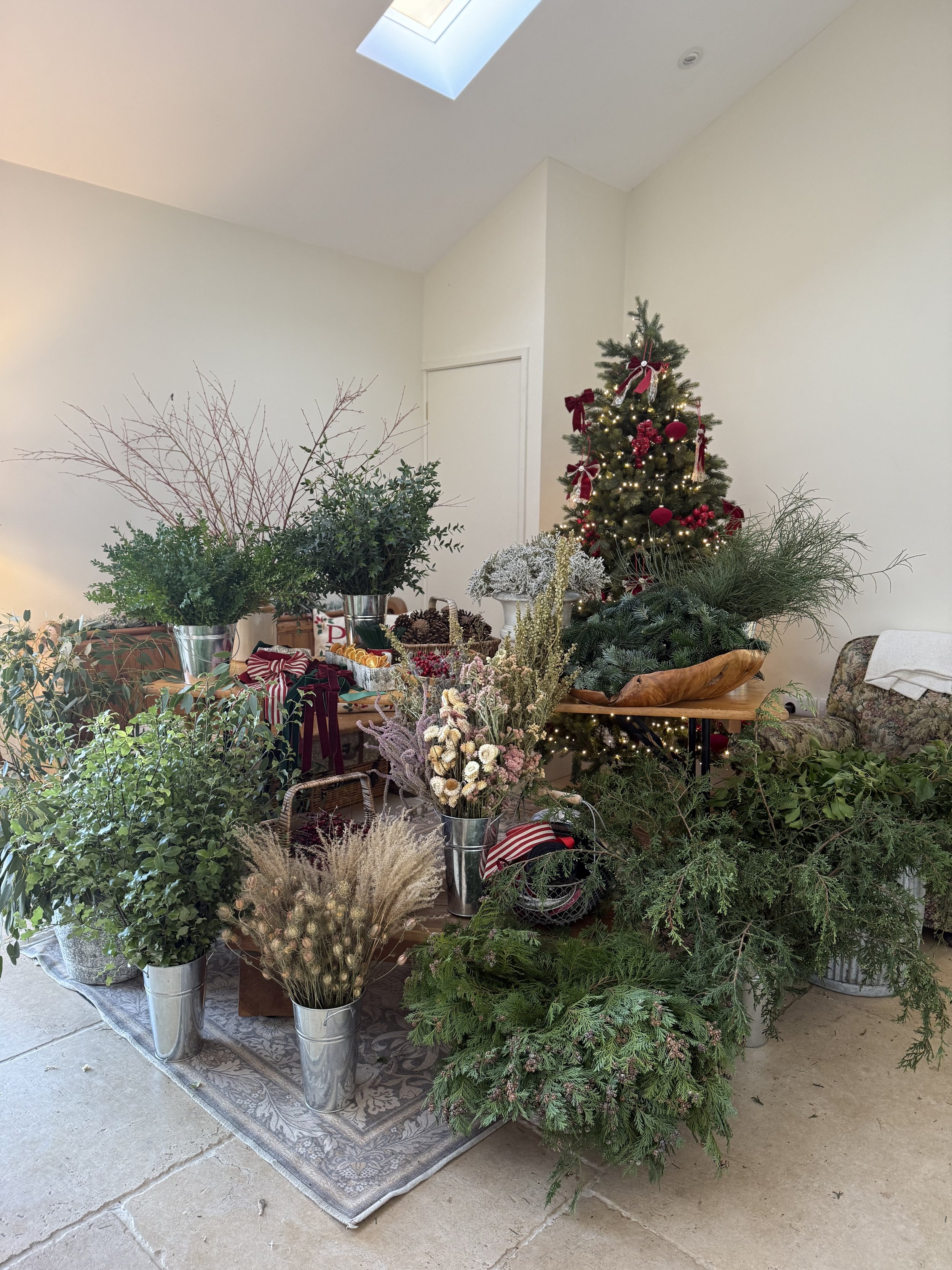 A Christmas tree decorated with red ornaments, ribbons, and lights in a bright room. Various potted plants and floral arrangements are displayed on tables and the floor nearby.
