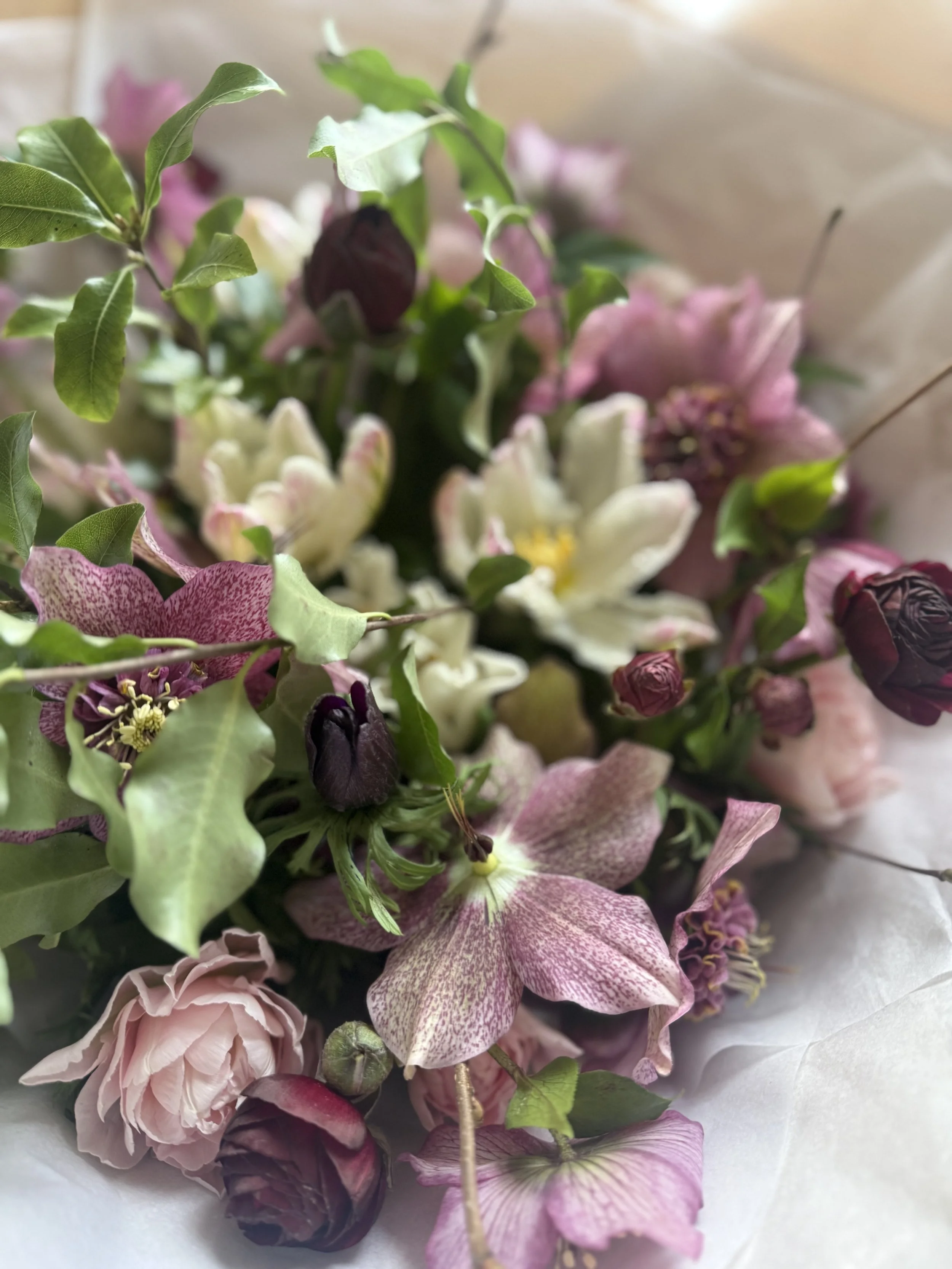 A close-up of a mixed bouquet of pink, white, and purple hellebore, tulip, ranunculus spring flowers with green leaves, wrapped in light-colored paper.