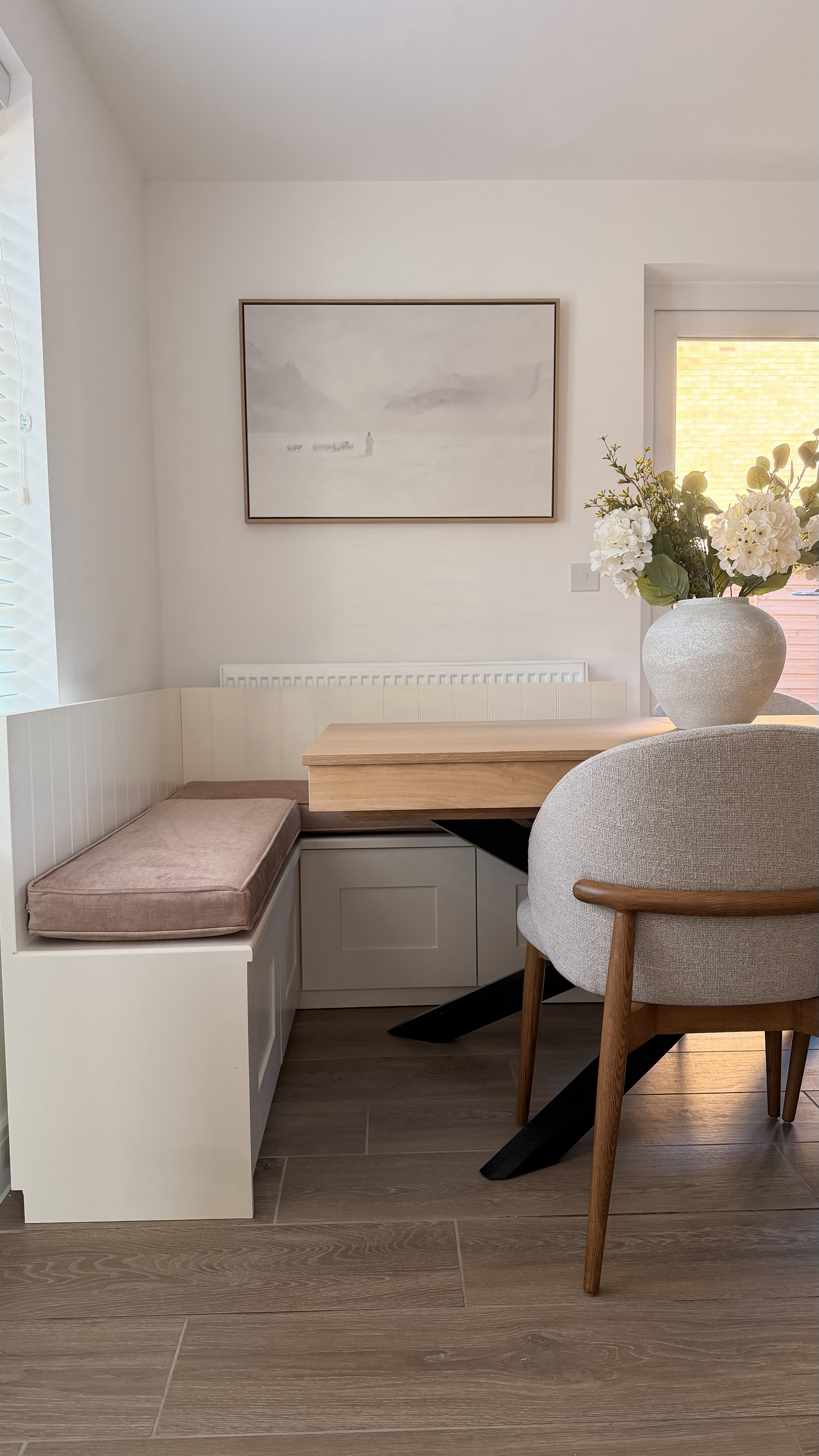 A cozy dining nook with built-in white seating with a pink cushion, a wooden table, and a beige upholstered chair with wooden legs. There is a large vase with white flowers on the table, a window with blinds letting in sunlight, a framed painting on 