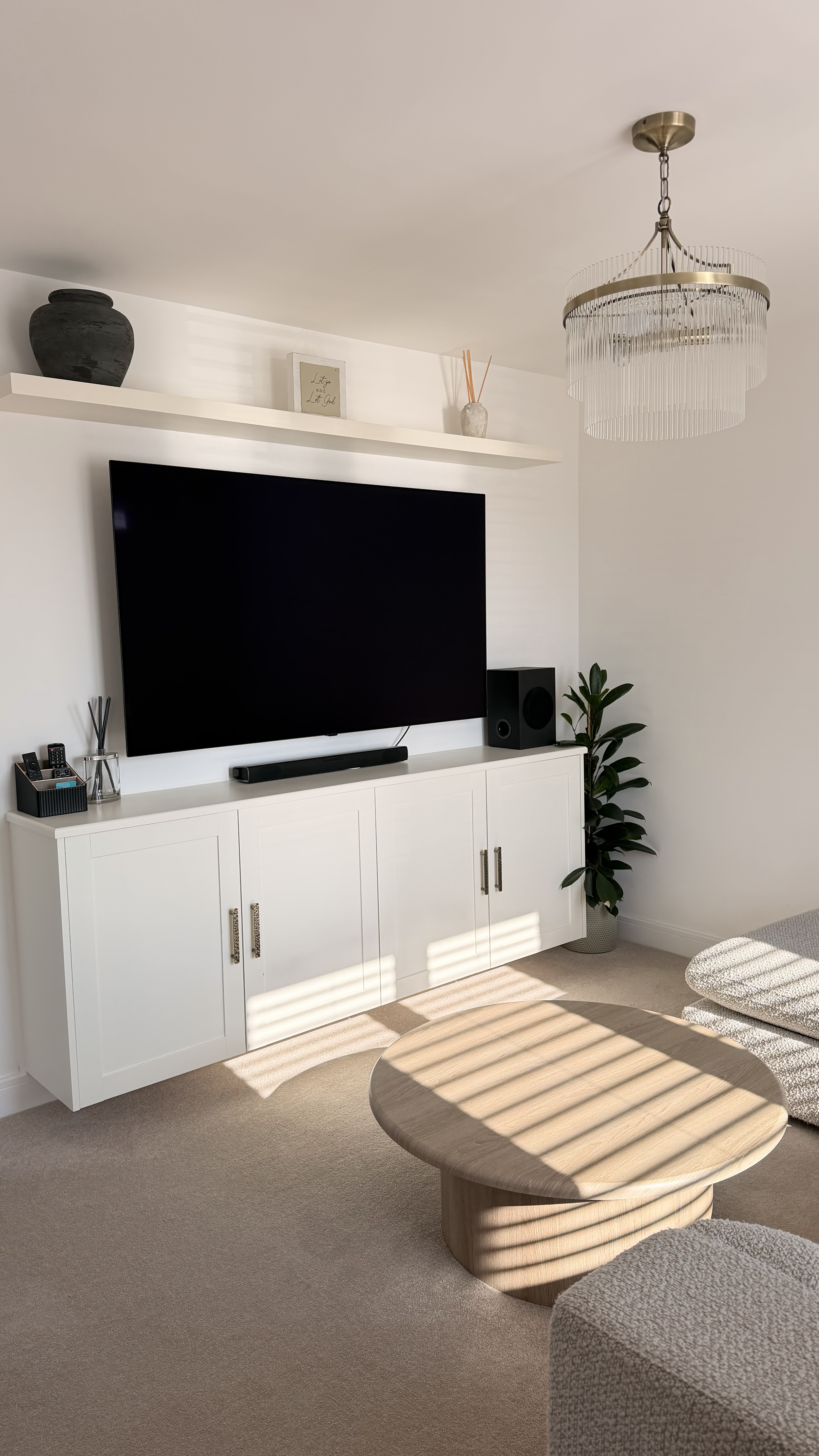 Living room with a flat-screen TV, a white entertainment cabinet, a potted plant, a wooden coffee table, and a modern chandelier. Sunlight casts shadows through window blinds.