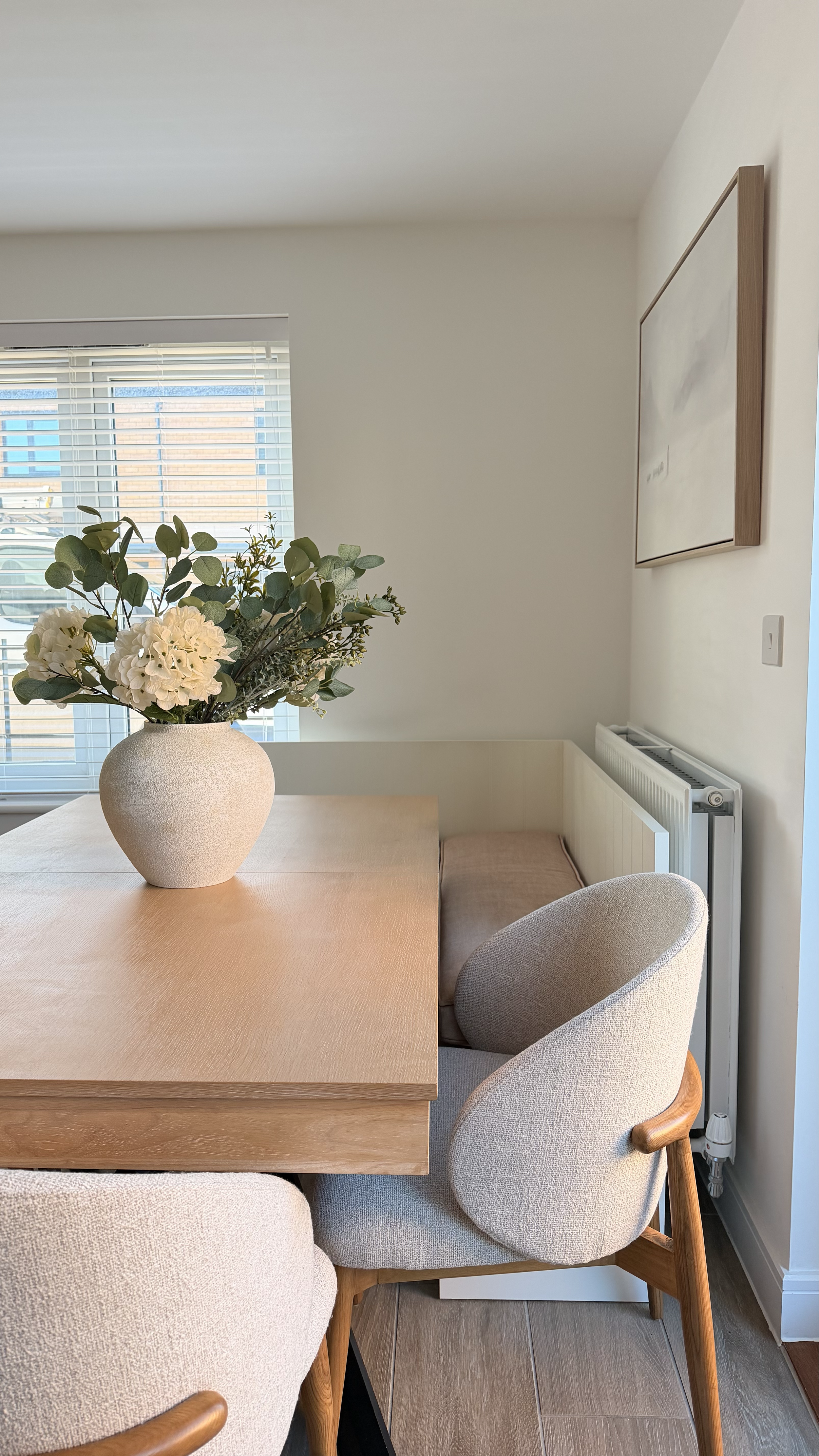 Dining room with a wooden table, beige upholstered chairs, a vase with white flowers on the table, and a window with blinds in the background.