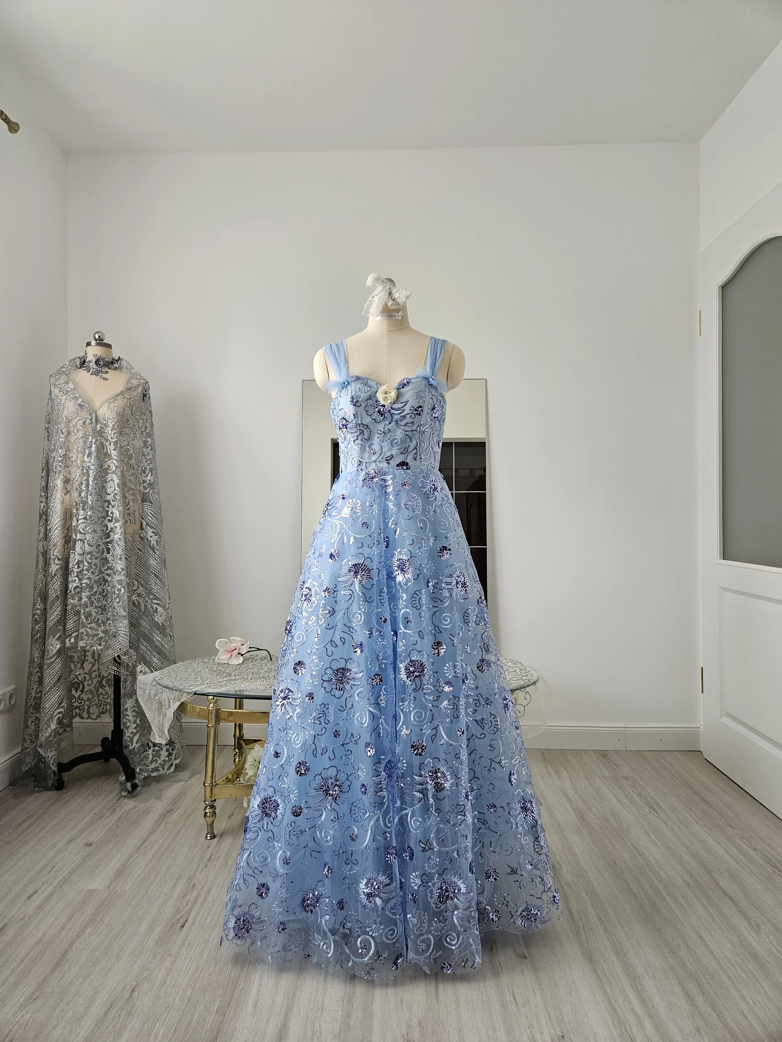 Abendkleid aus eisblauem Tüll mit floralen Glitzerpailletten, Herzausschnitt und breiten Trägern. Es hat eine weiße Zierblüte und Perlen und Strass.