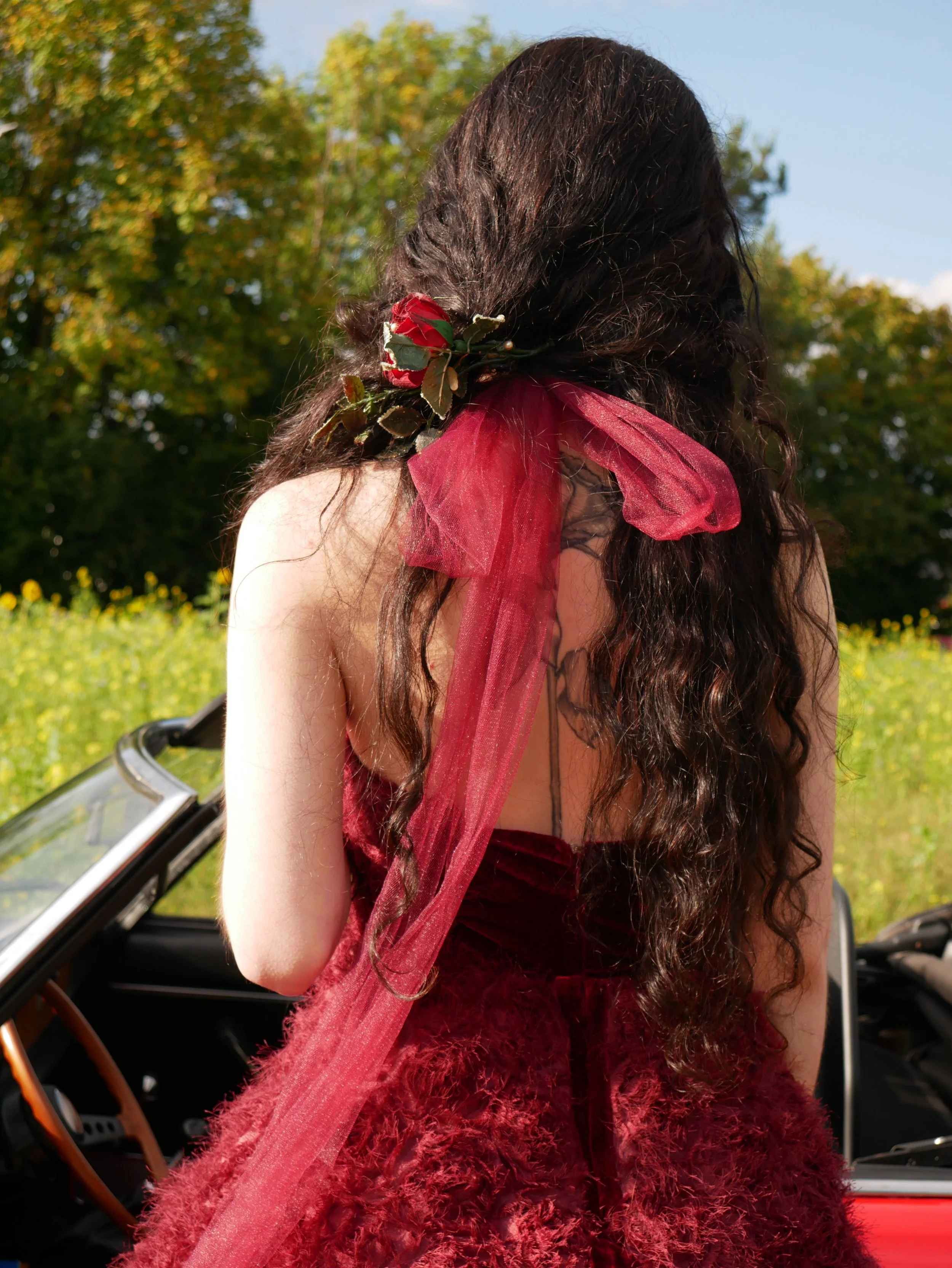 Eine Frau von hinten, in einem dunkelroten Rosenkleid mit breiten Neckholdern. Ihr langes, schwarzes Haar wird mit einer einer Rose geschmückt.