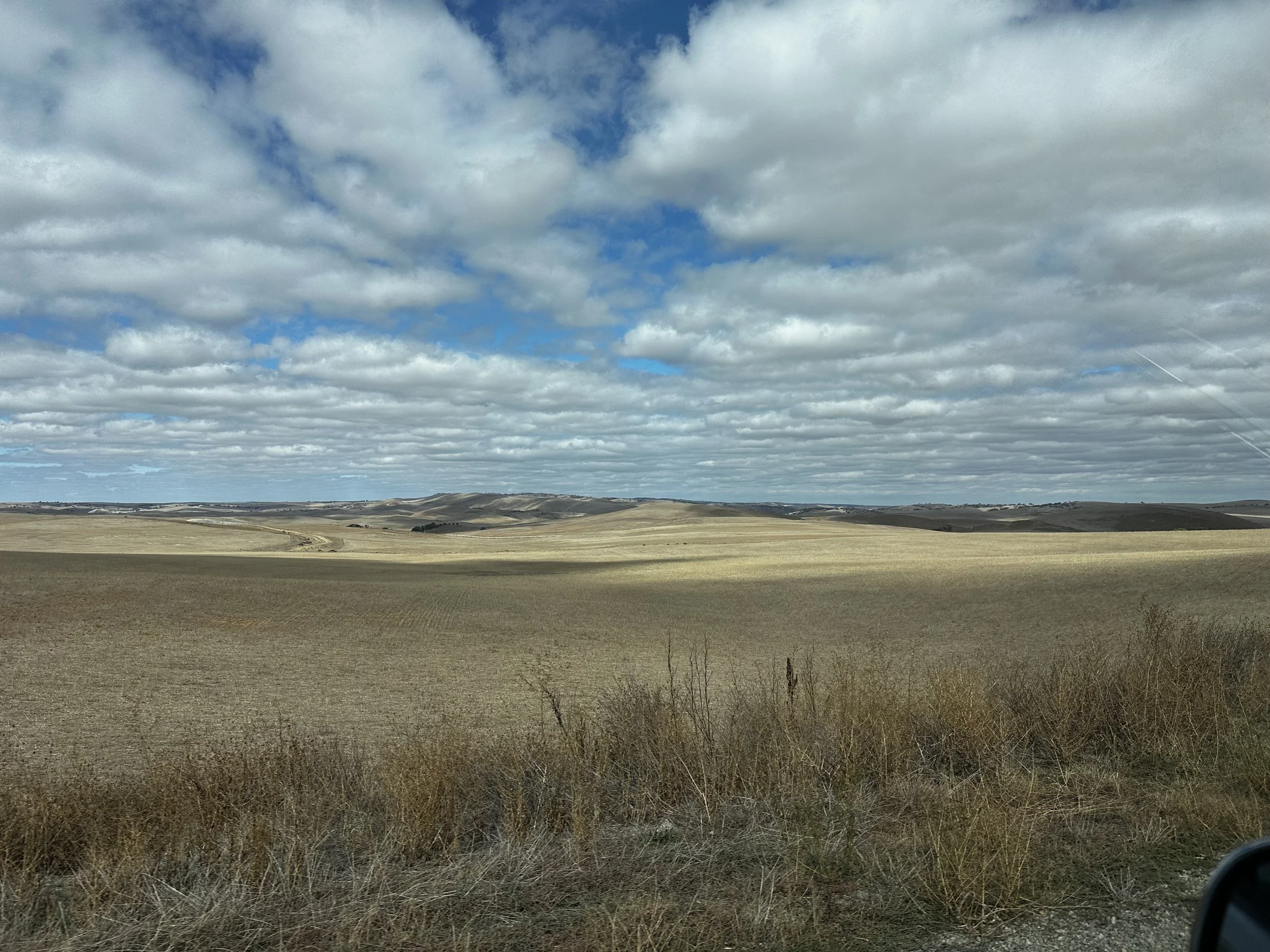 Open landscape with rolling hills, brown and green fields, and a vast sky filled with scattered clouds.