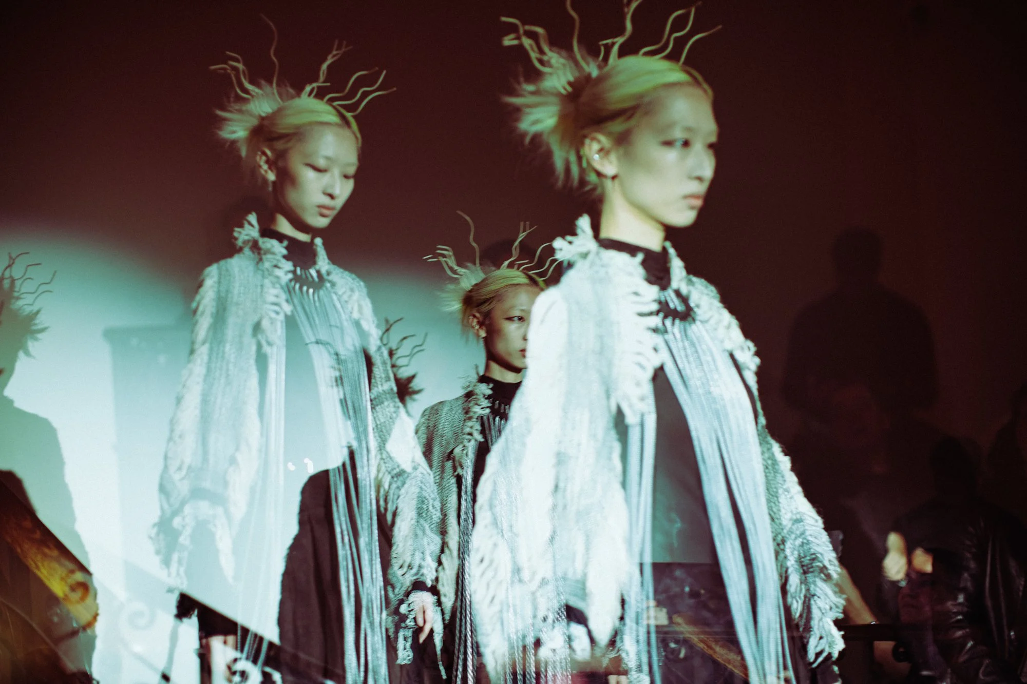 Multiple women in elaborate fashion outfits with flowing patterns, standing on a runway during a fashion show, reflected on a glass surface.