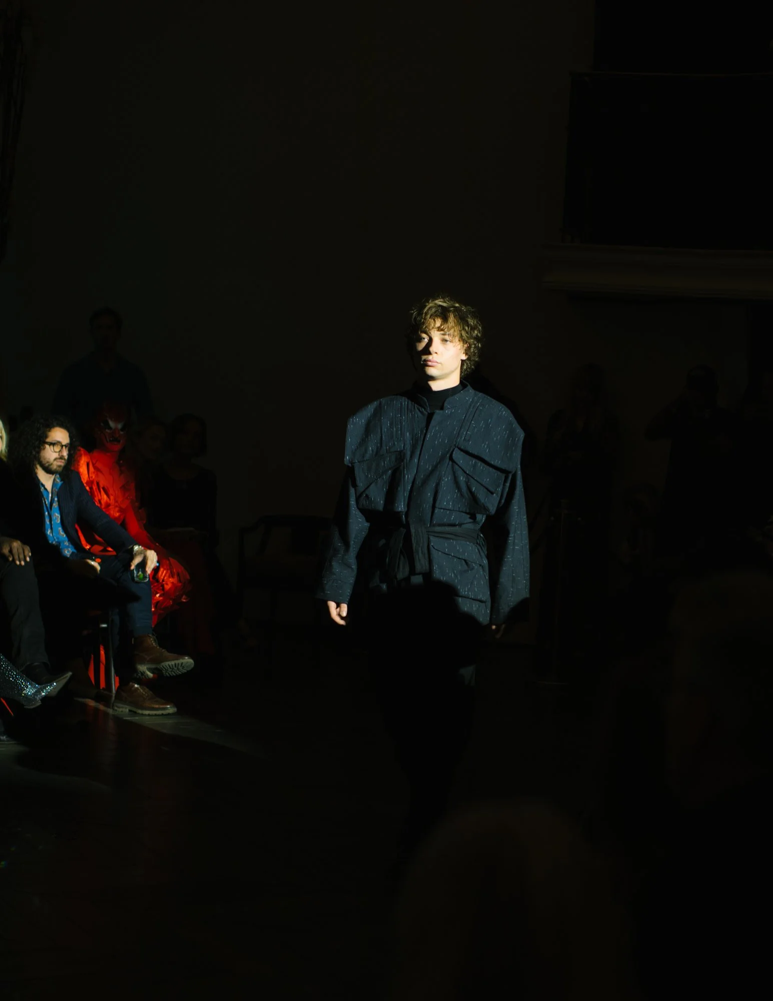 A male model walking on a fashion runway with spotlights, audience seated on sides, dark background.