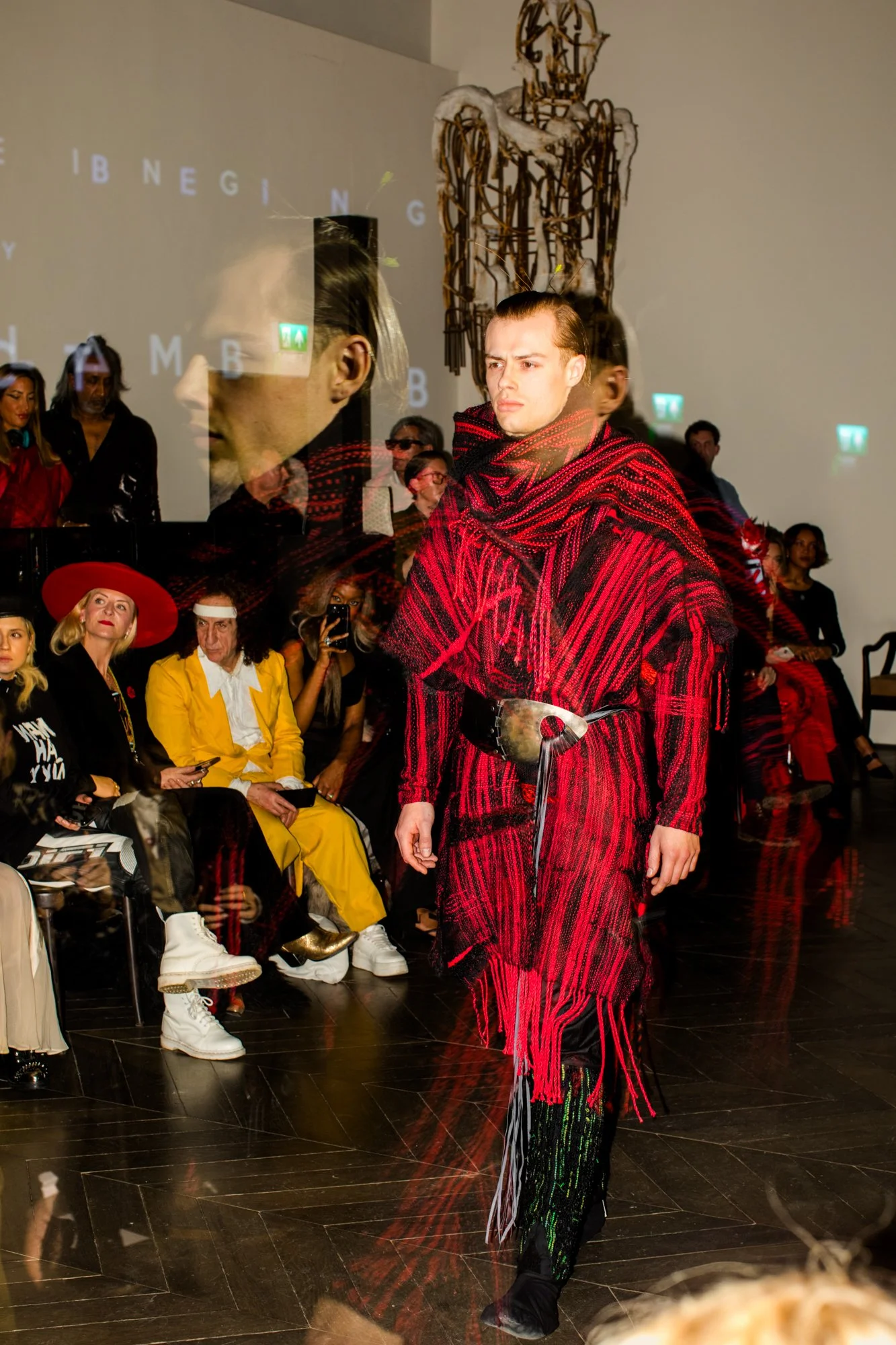 Model walking down runway at a fashion show, wearing a red and black striped outfit with a wide belt, in front of seated audience.