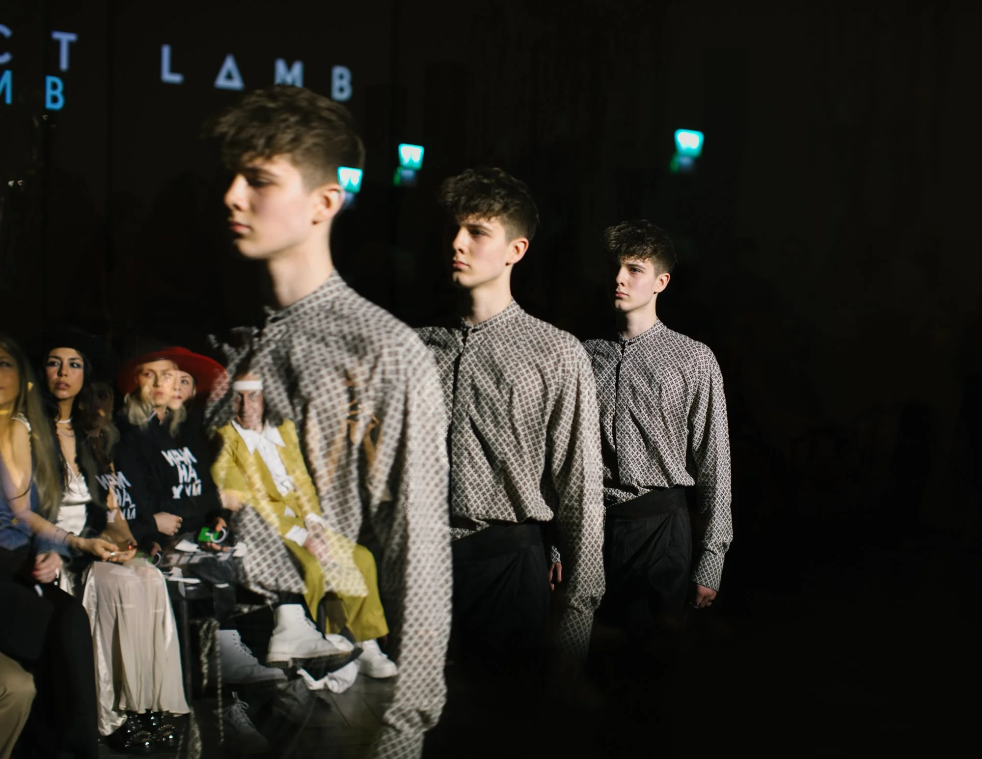 Models walking on a runway during a fashion show, with spectators seated on the side.