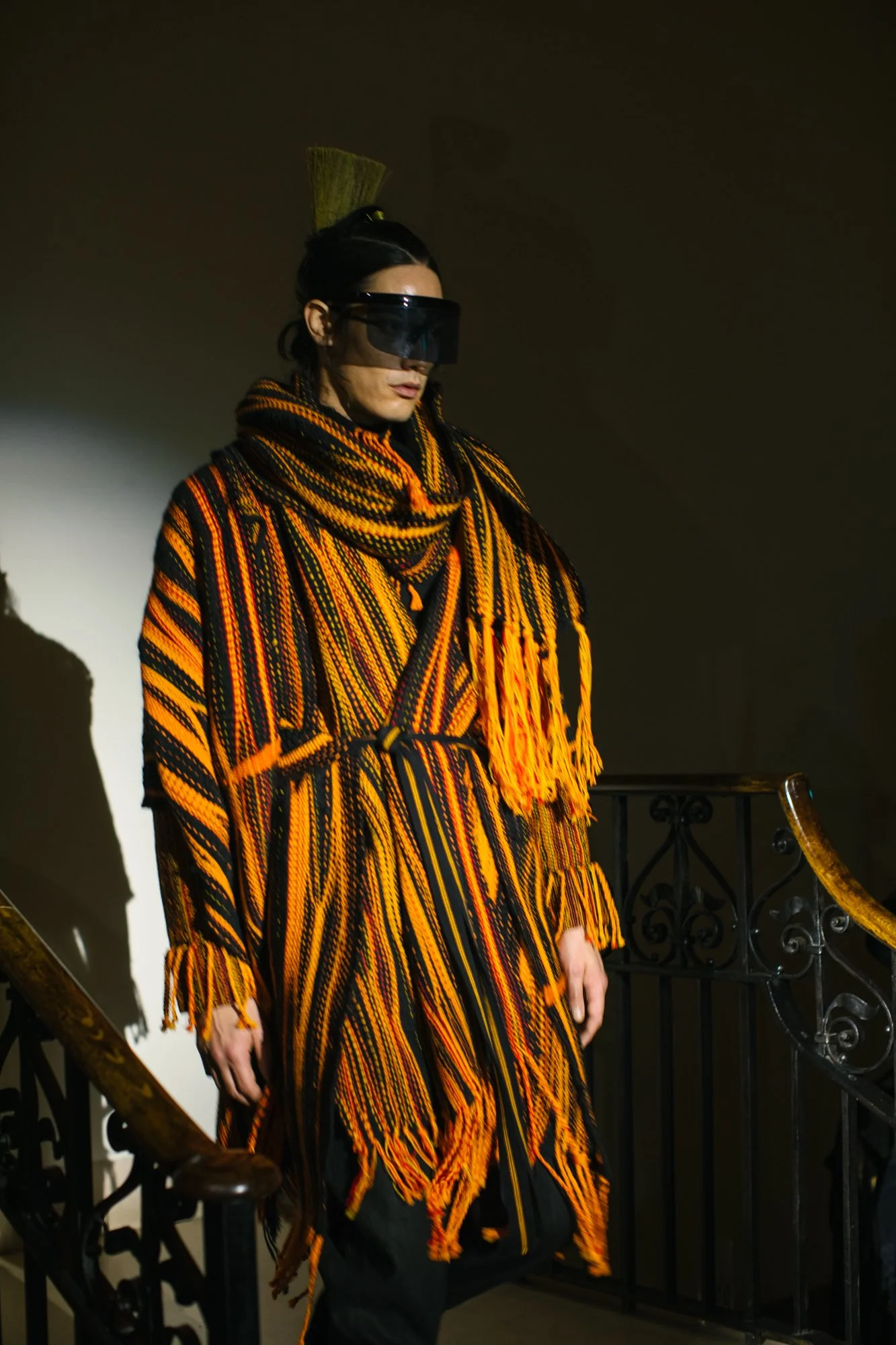 Person wearing a striped, fringed orange and black coat, black pants, and futuristic visor, standing on a staircase with a dark background.