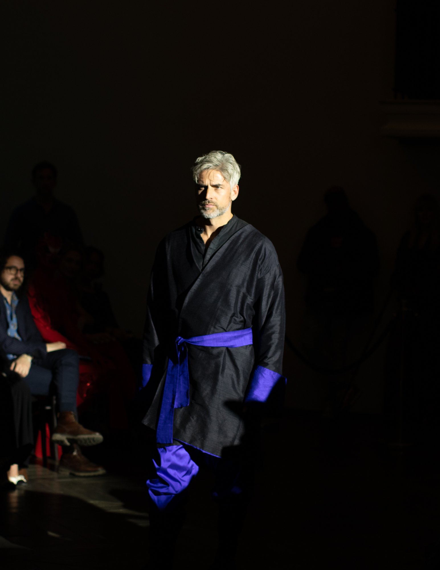 A man with gray hair and beard walking on a runway during a fashion show, wearing a black and purple outfit.