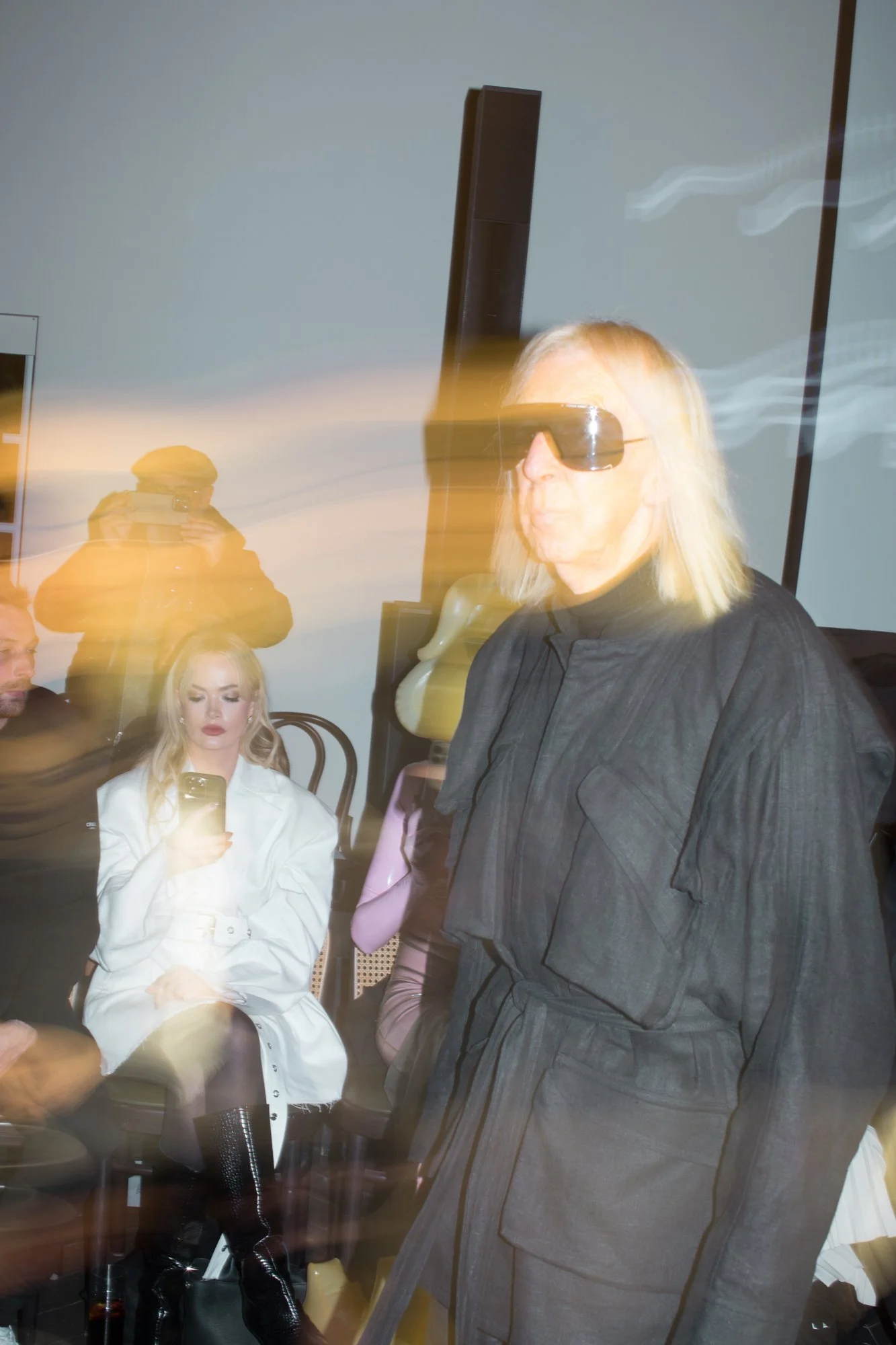 A woman with blonde hair wearing a white coat sitting and looking at her phone, and an older woman with blonde hair and sunglasses standing nearby, in a room with blurry streaks of light.