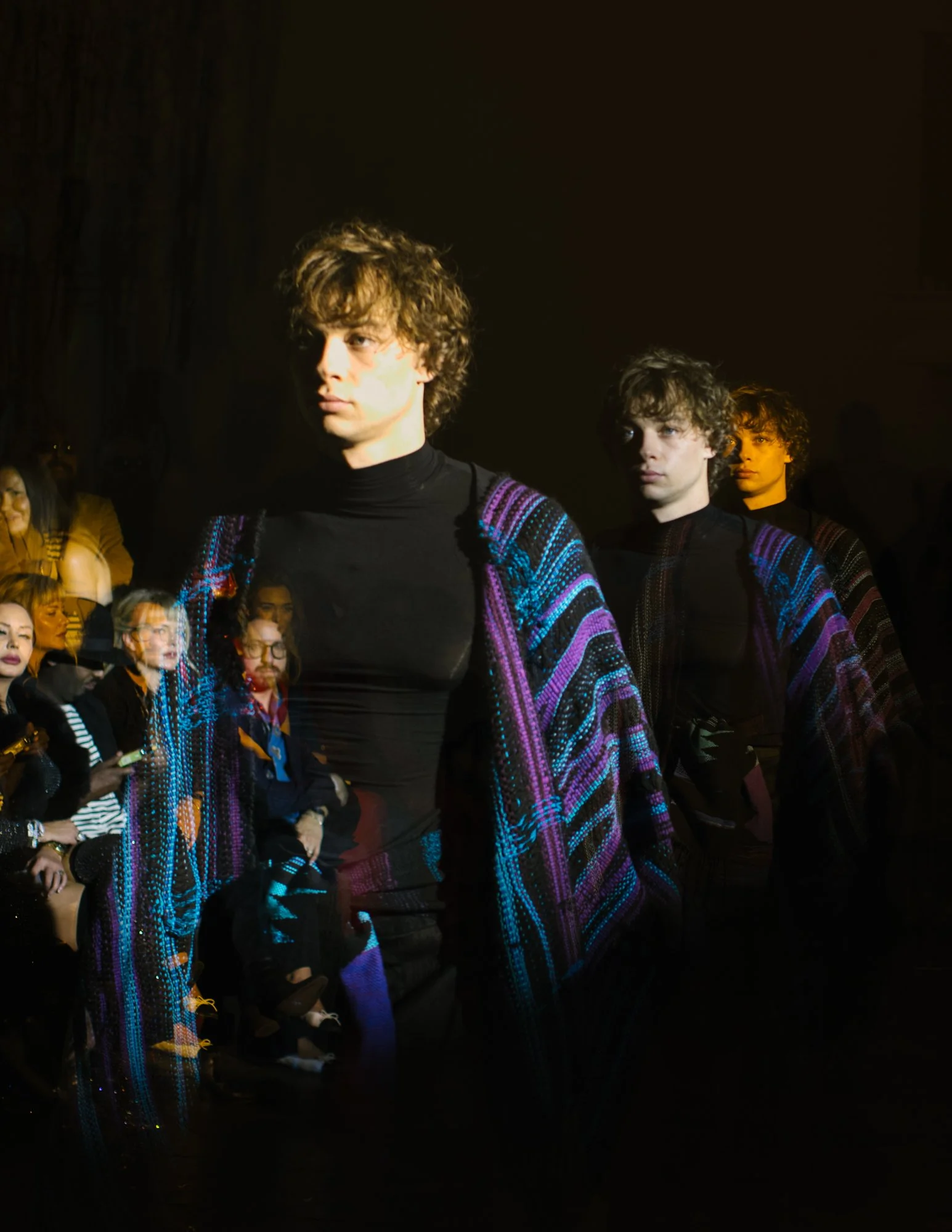 Models walking down the runway during a fashion show, wearing colorful, patterned garments. Audience seated on either side of the runway watching the show.