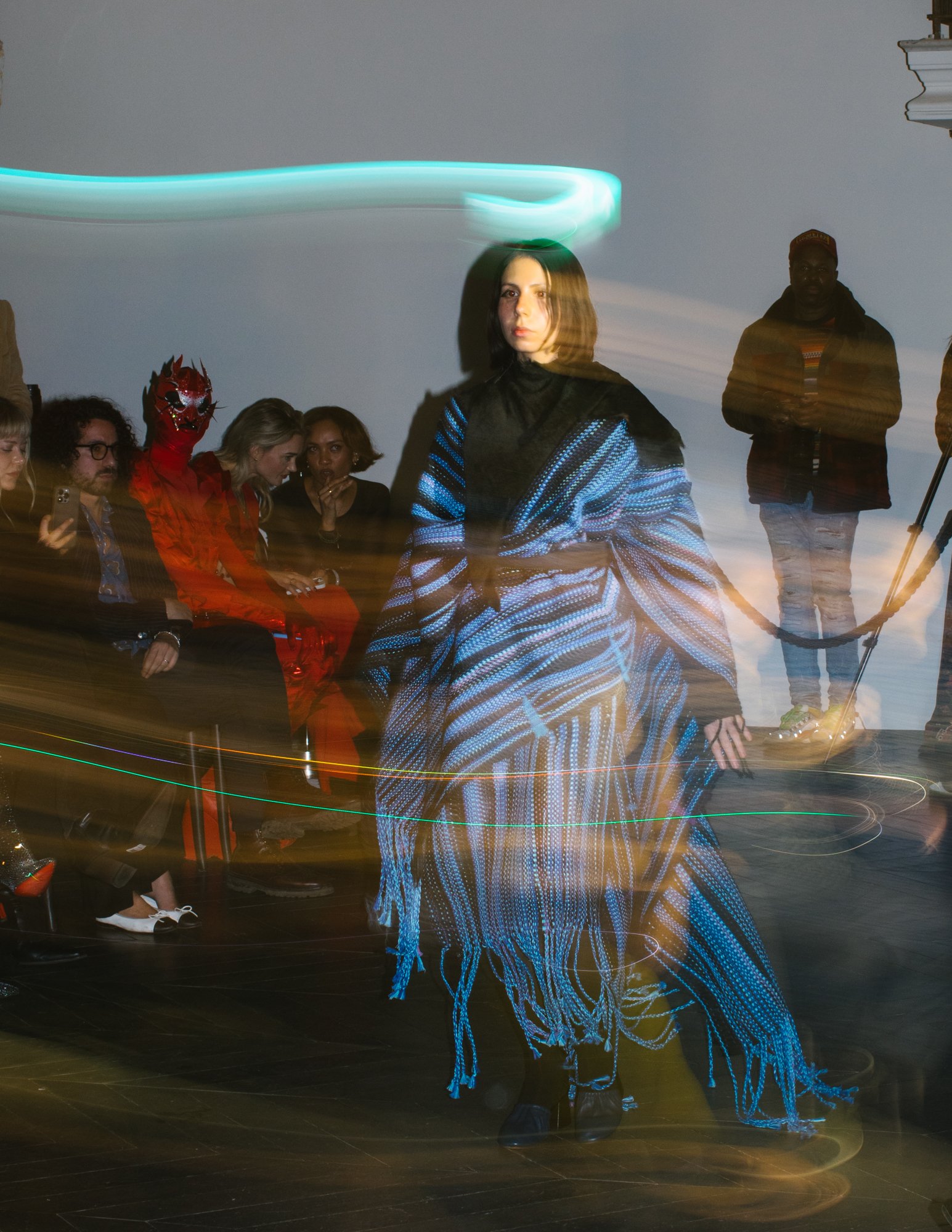 A woman walking on a runway during a fashion show, wearing a striking blue and black patterned outfit. The audience is seated on either side of the runway, some taking photos. Lights create motion streaks around her.