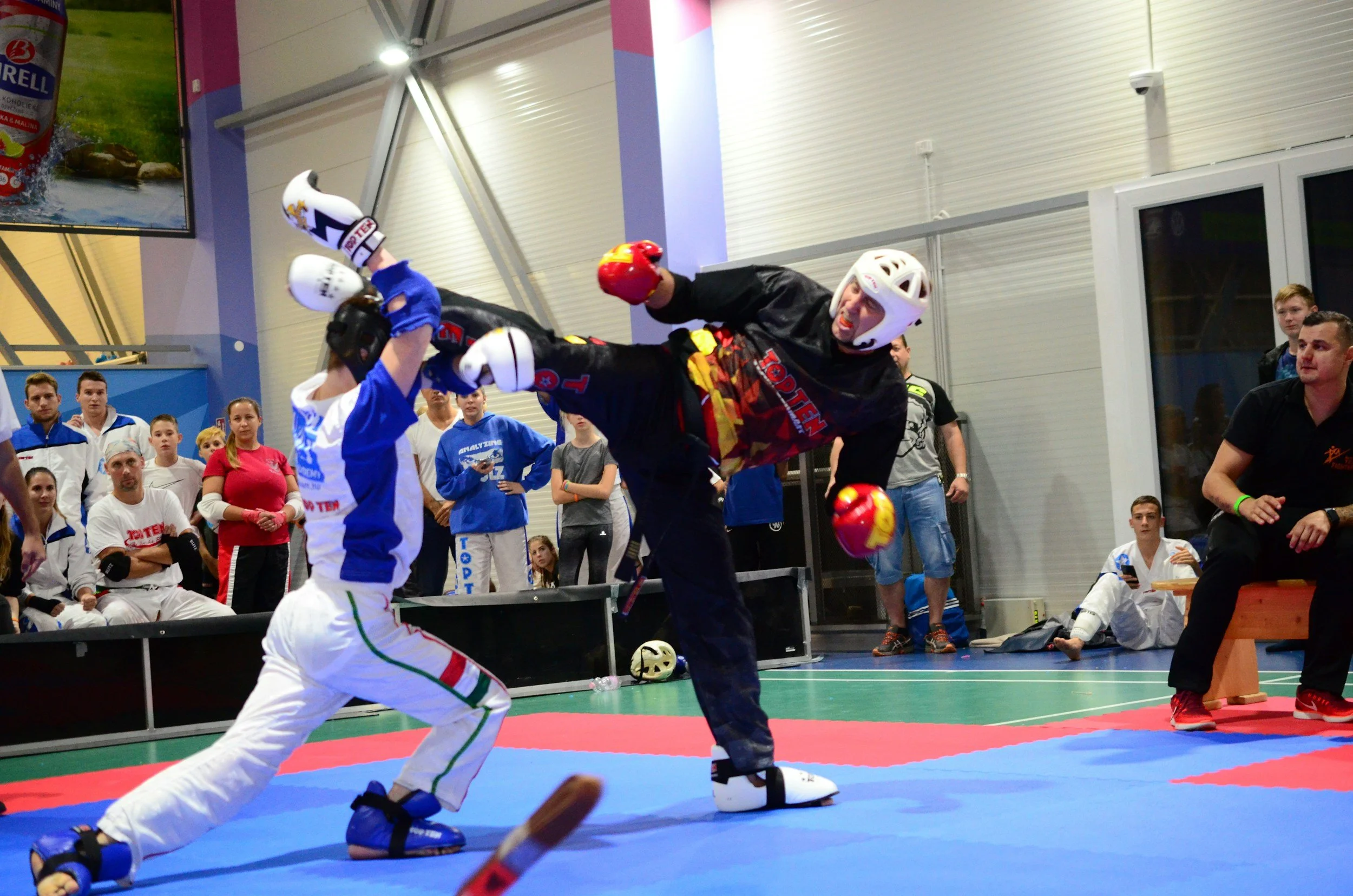 Two martial artists engaged in a match on a blue and red mat, wearing protective gear including helmets and gloves, with a group of spectators watching in the background at an indoor sports venue.