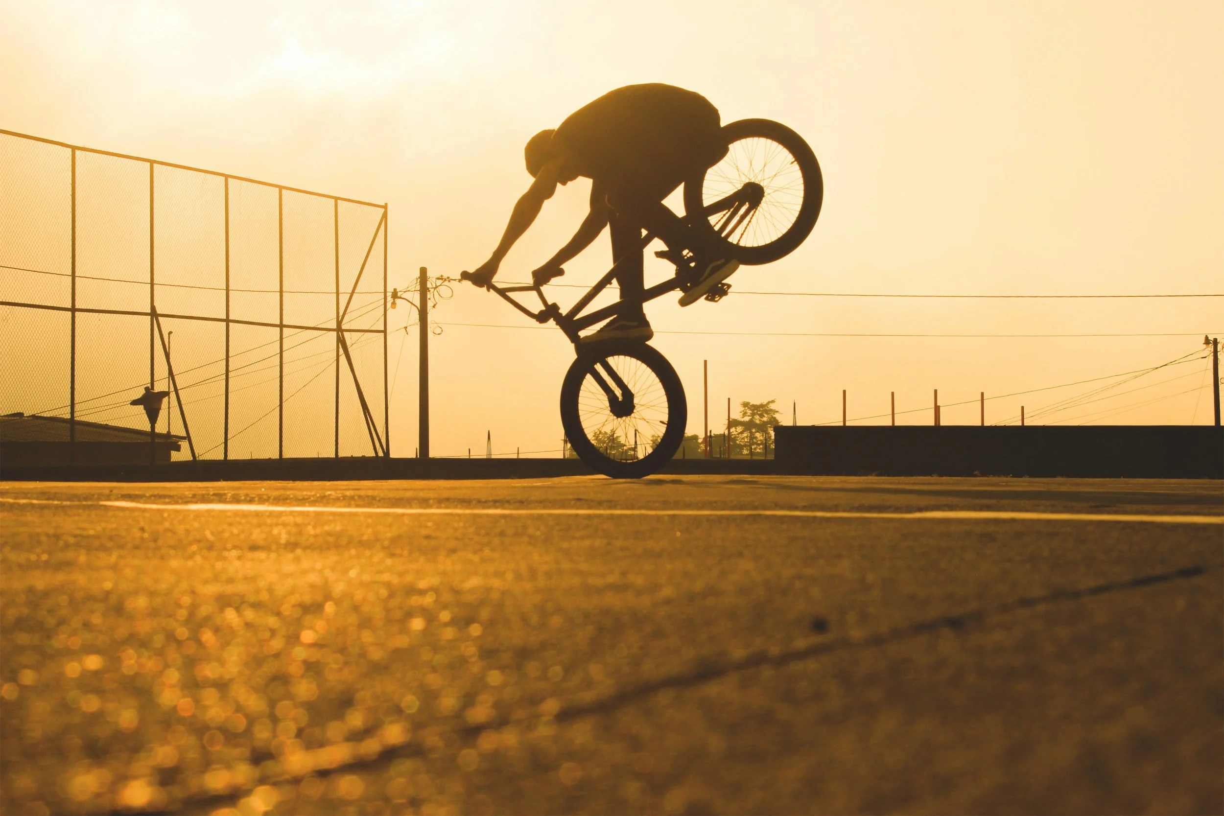 Person performing a BMX trick at sunset, mid-air with a bike upside down, in a sports area with a fence and power lines.