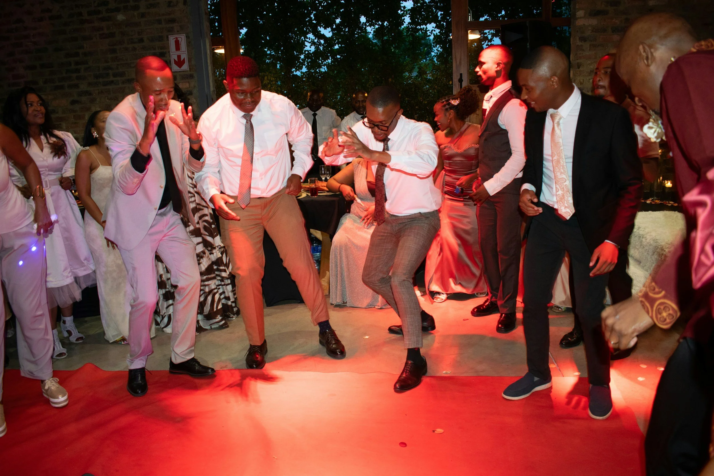 People dancing on a red carpet at a celebratory event in a dimly lit room with brick walls and string lights.