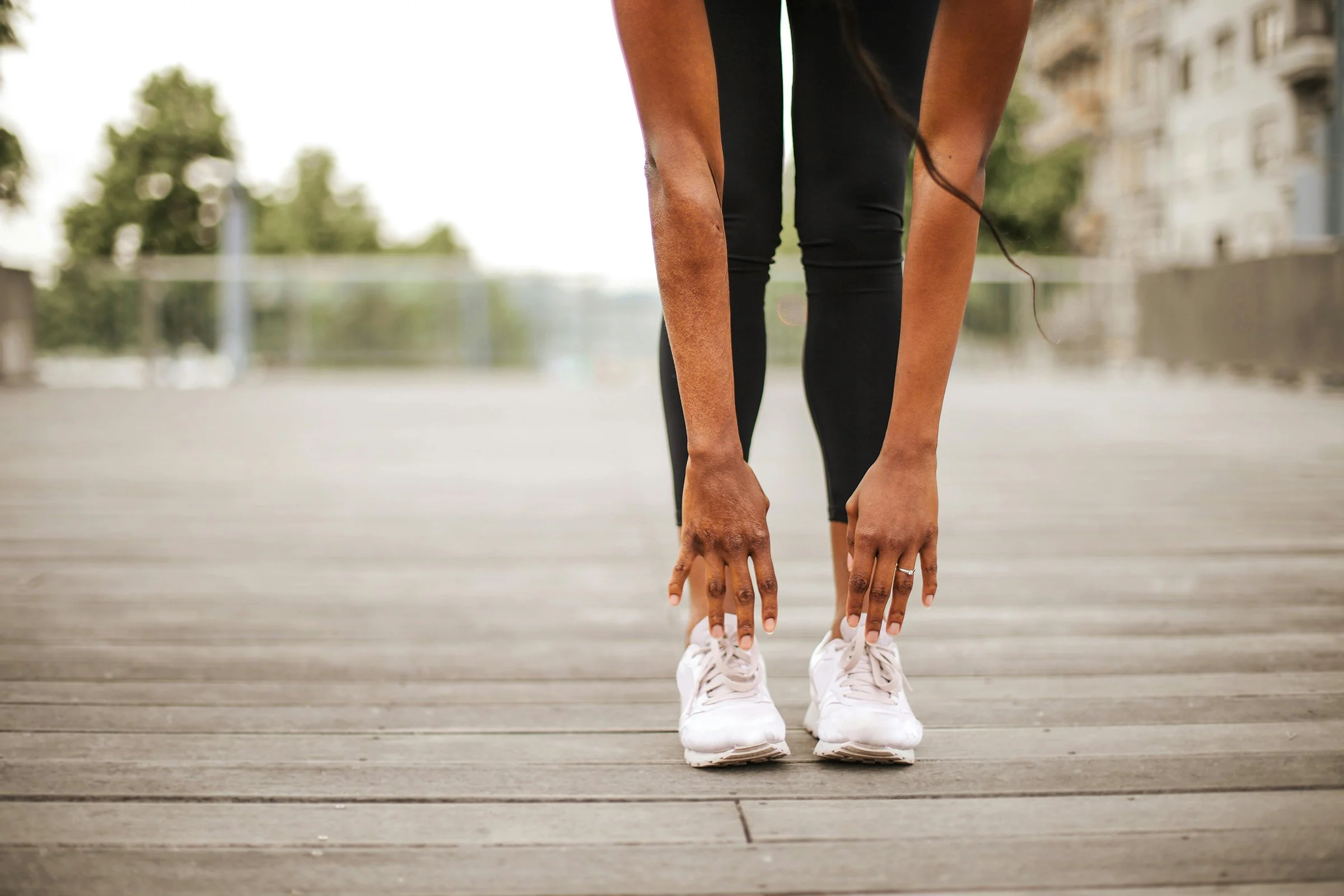 Person in black leggings and white running shoes stretching outdoors on a wooden bridge or dock.
