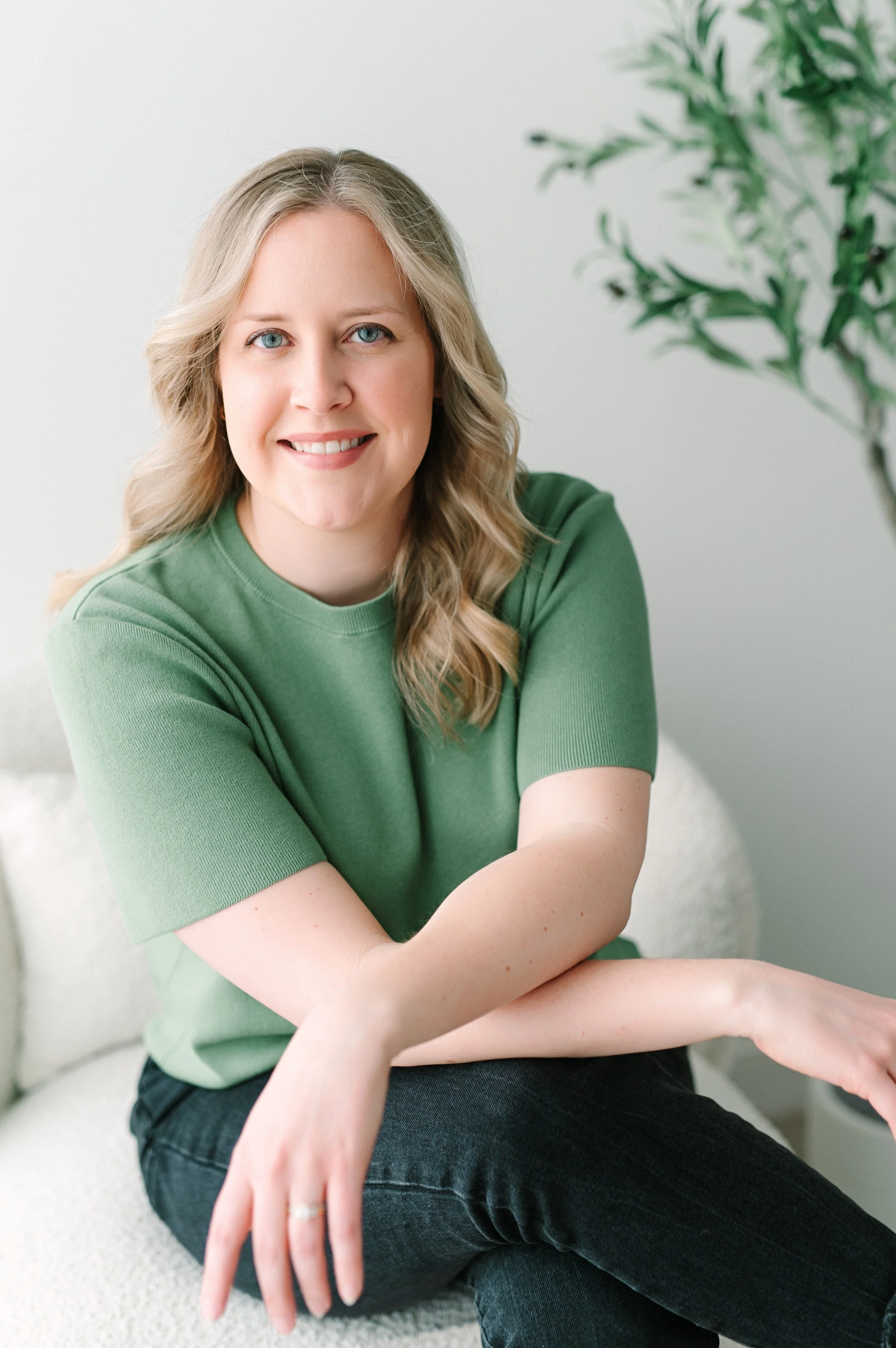 A woman smiling with blonde wavy hair, wearing a green shirt, sitting indoors near a potted plant with green leaves.