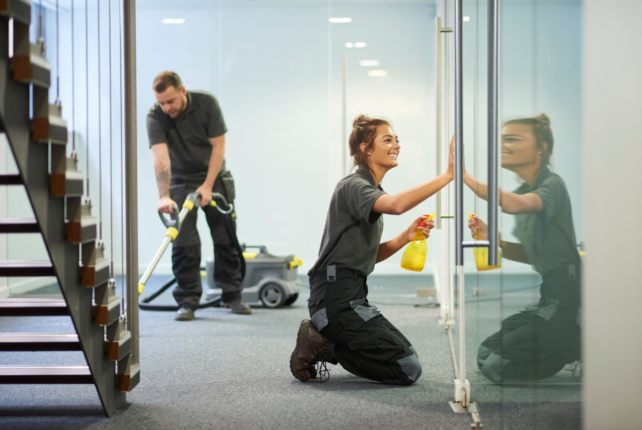 Two janitors cleaning an office; a woman cleaning glass with a spray bottle and a man vacuuming the carpet.