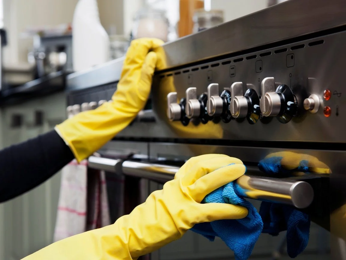 Person wearing yellow rubber gloves cleaning an oven with a blue cloth in a kitchen.