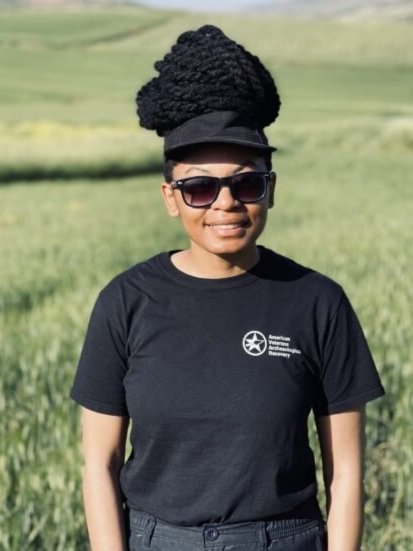Woman wearing sunglasses and a black T-shirt with an American Cancer Society logo, standing outdoors in a grassy field with hills in the background.