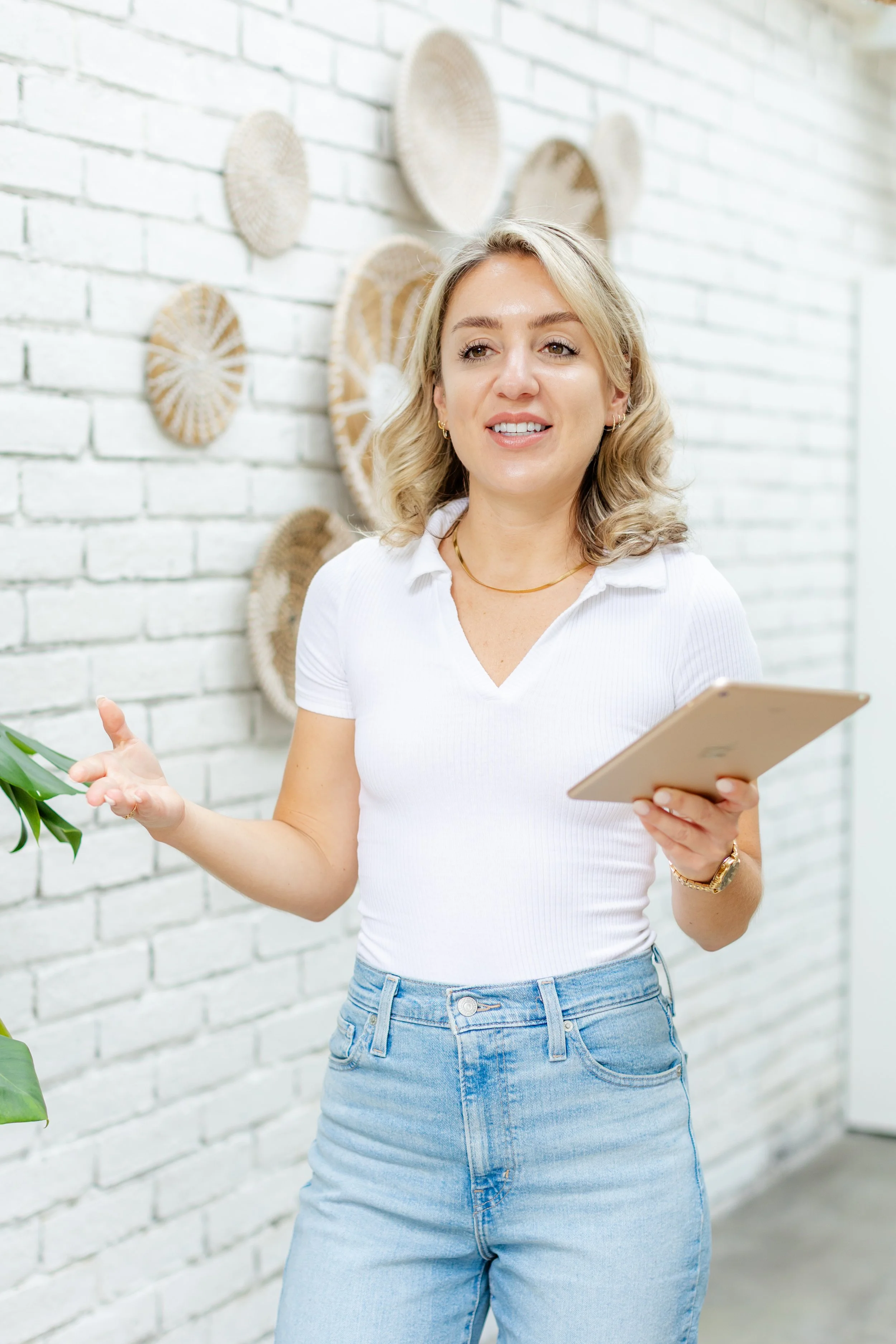 A woman in a white shirt and jeans holding a tablet while speaking, standing in front of a white brick wall decorated with woven wall hangings.