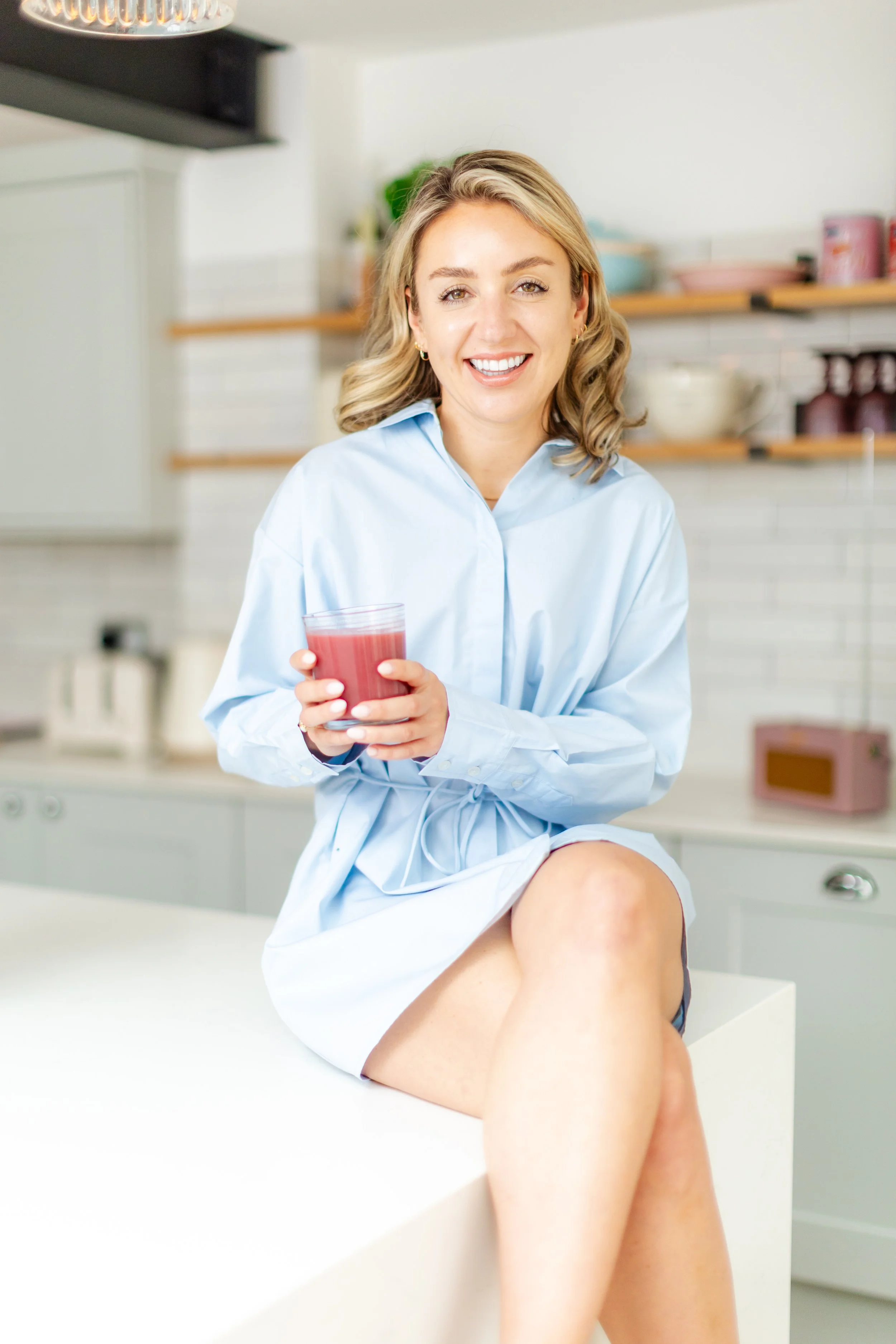 A woman with blonde hair wearing a light blue shirt dress, sitting on a kitchen counter, smiling, holding a glass of pink juice.