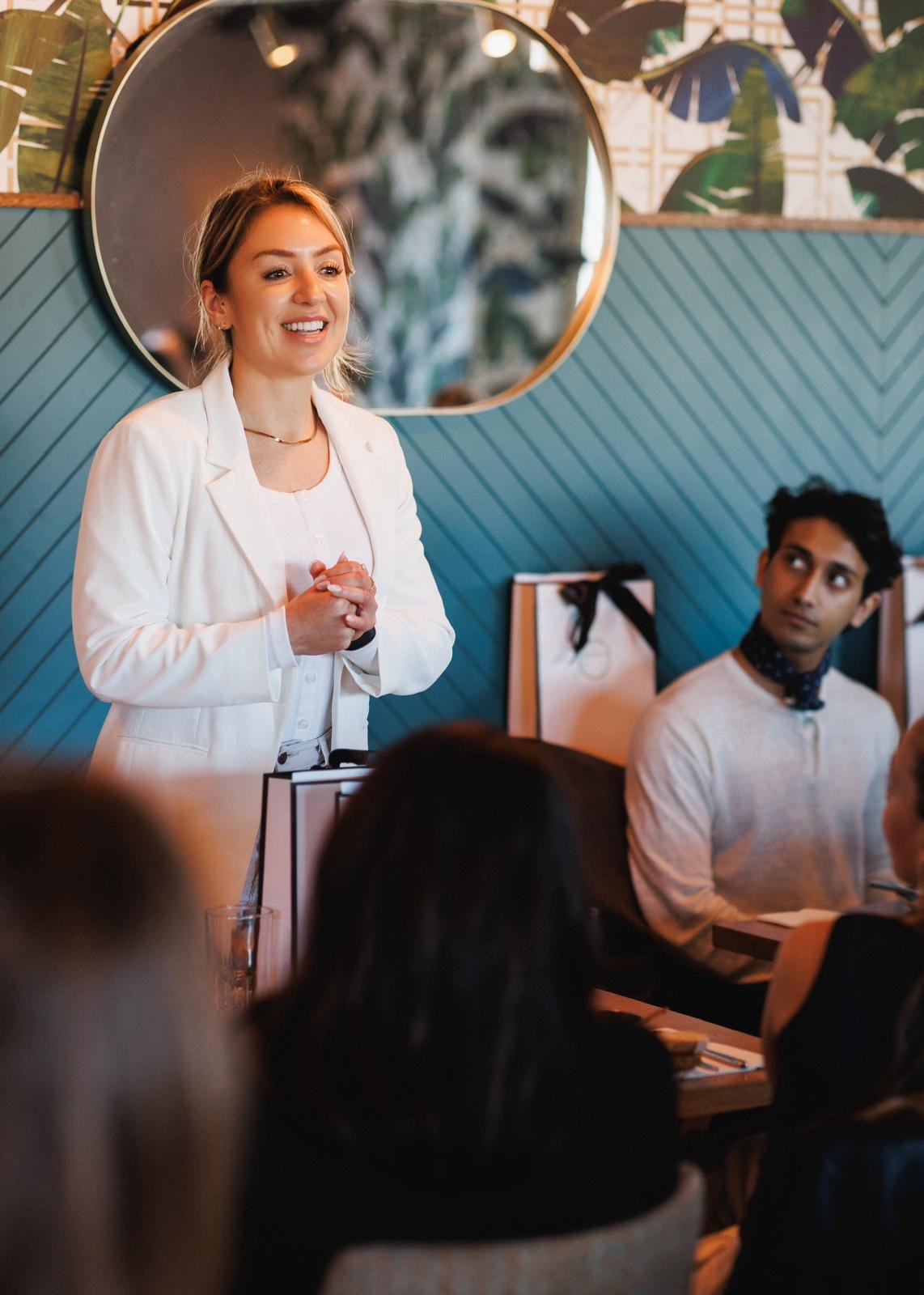A woman in a white blazer speaking to an audience in a room with blue and tropical-themed wallpaper, while a man in a white shirt and neck scarf listens, seated at a table.
