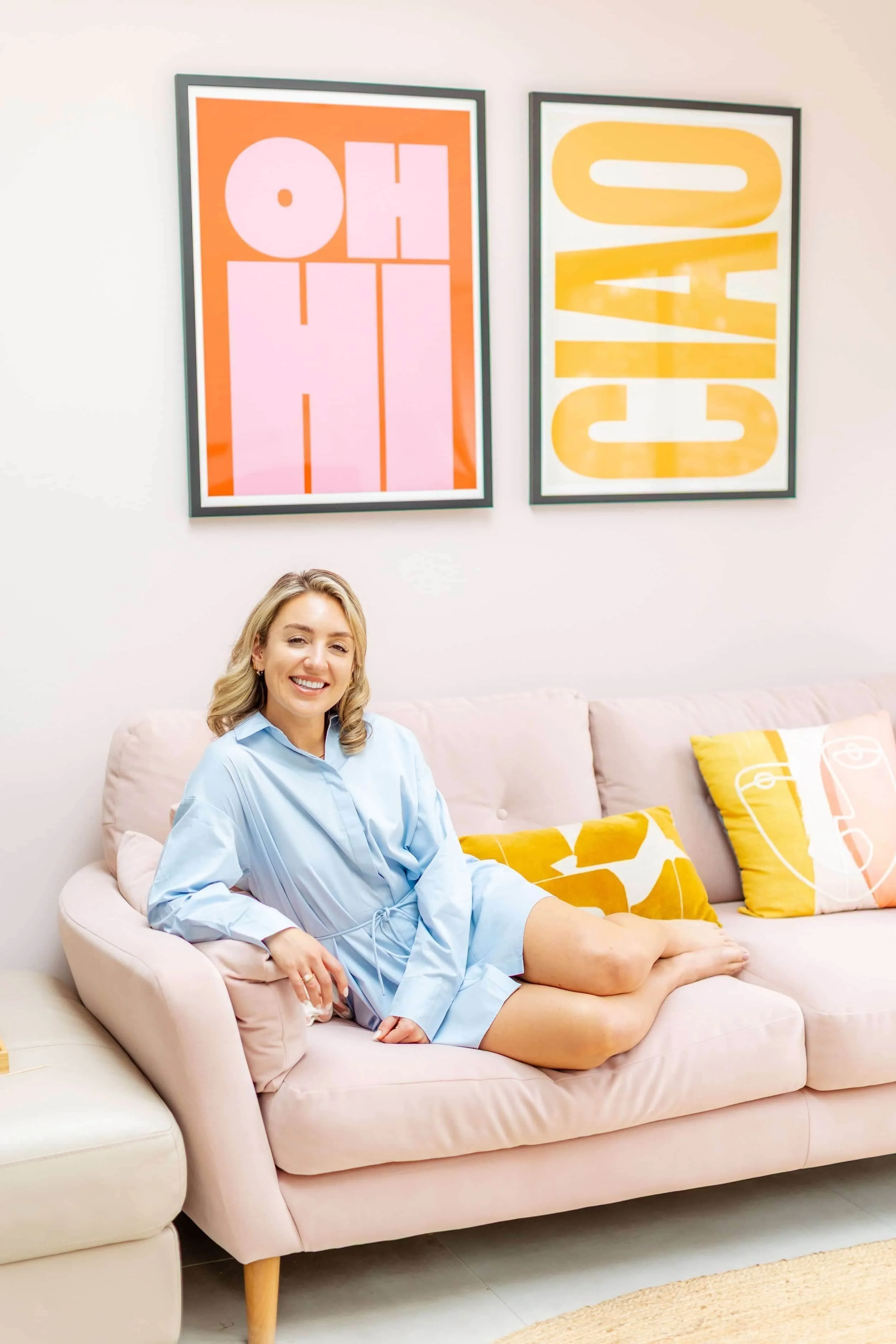 A young woman with blonde hair sits on a pink couch, smiling at the camera. She is wearing a light blue robe, with her legs crossed. Behind her are two framed artworks with bold, colorful text that read 'OH' and 'COACH'. The couch has yellow pillows with orange and white face illustrations.