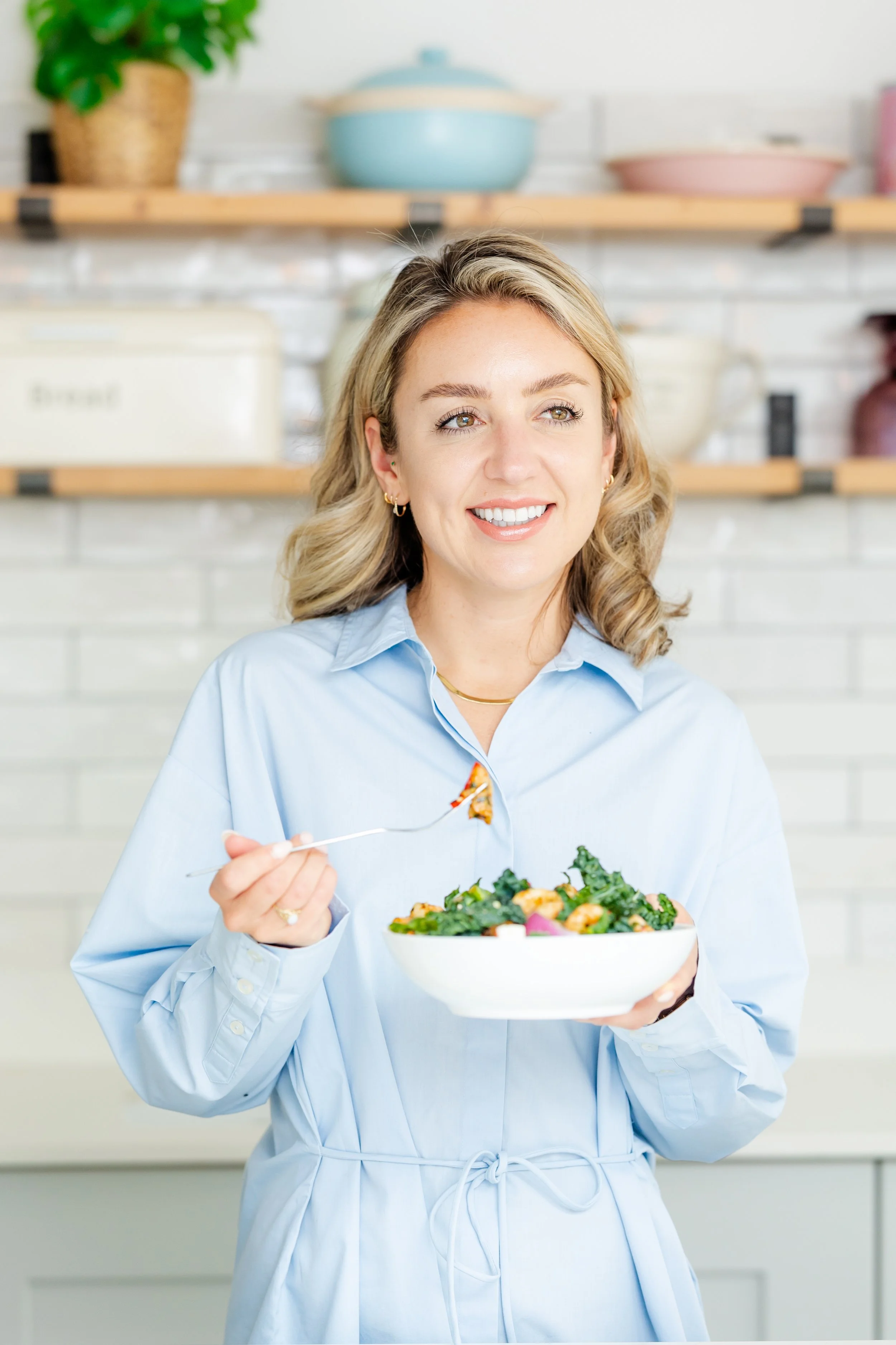 A woman with blonde hair in a blue shirt holding a bowl of salad and a fork, standing in a bright kitchen.