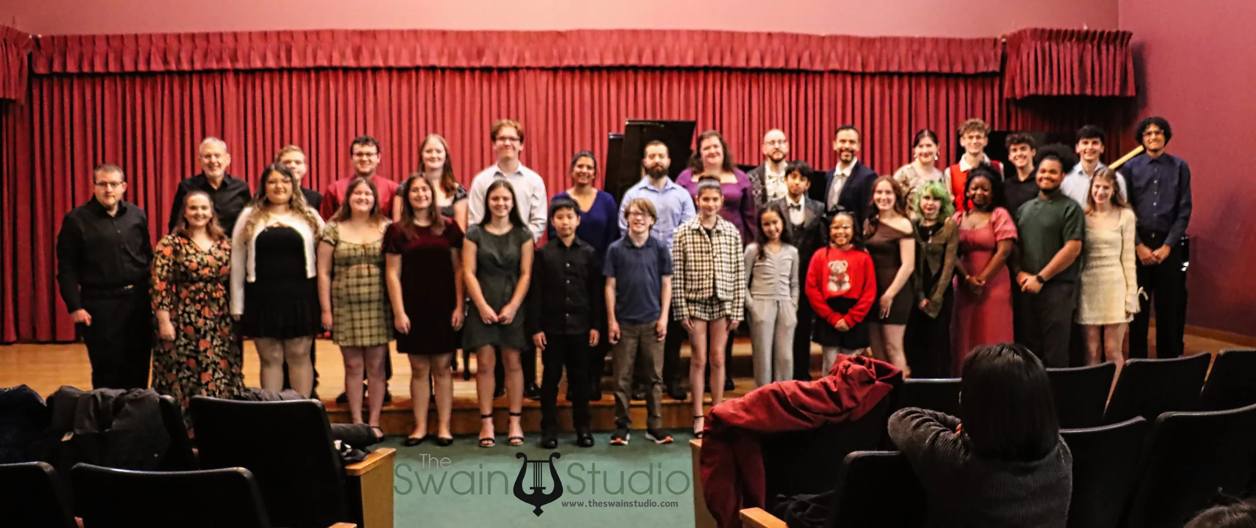 A large group of people, including children and adults, poses on a stage in front of a red curtain, likely after a performance or event. Some are smiling, and a few are dressed in festive or formal attire.