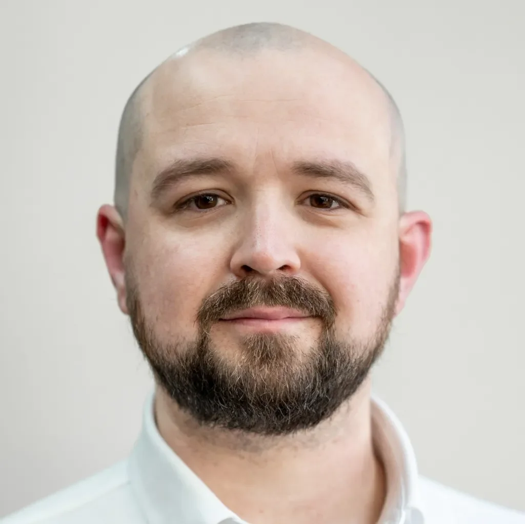 Man with brown eyes, shaved head and a beard smiling softly at the camera