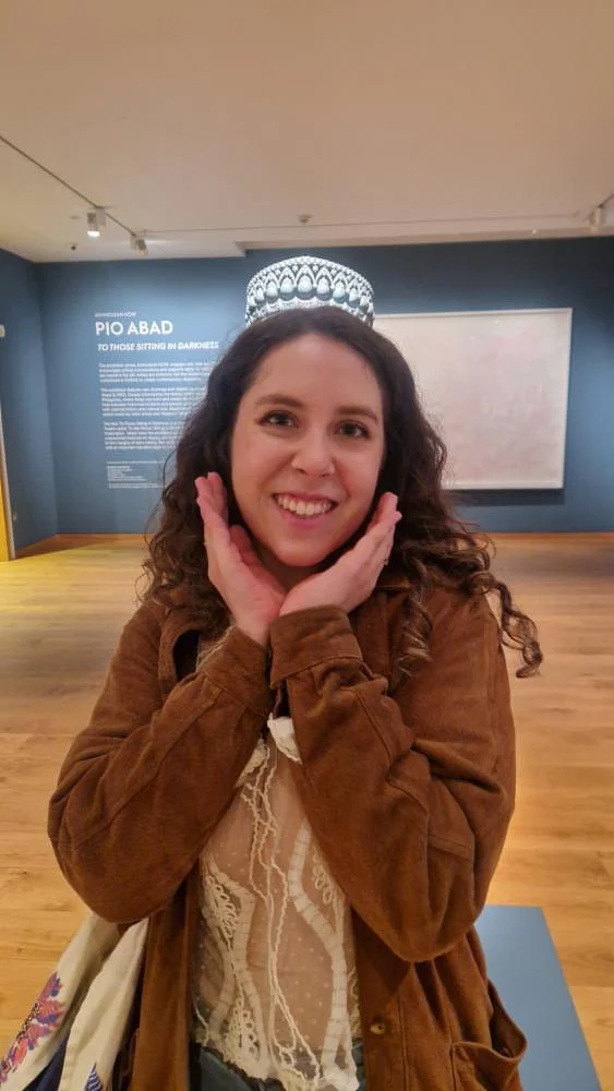 A smiling woman with curly hair wearing a brown jacket and a lace top, posing with her hands on her cheeks in an art gallery.