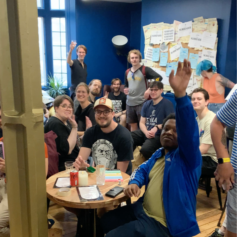 Group of people gather in a room with blue walls, some sitting at a round table with drinks, others standing or sitting in the background. One person in the foreground raises their hand, and others are smiling and enjoying the moment.