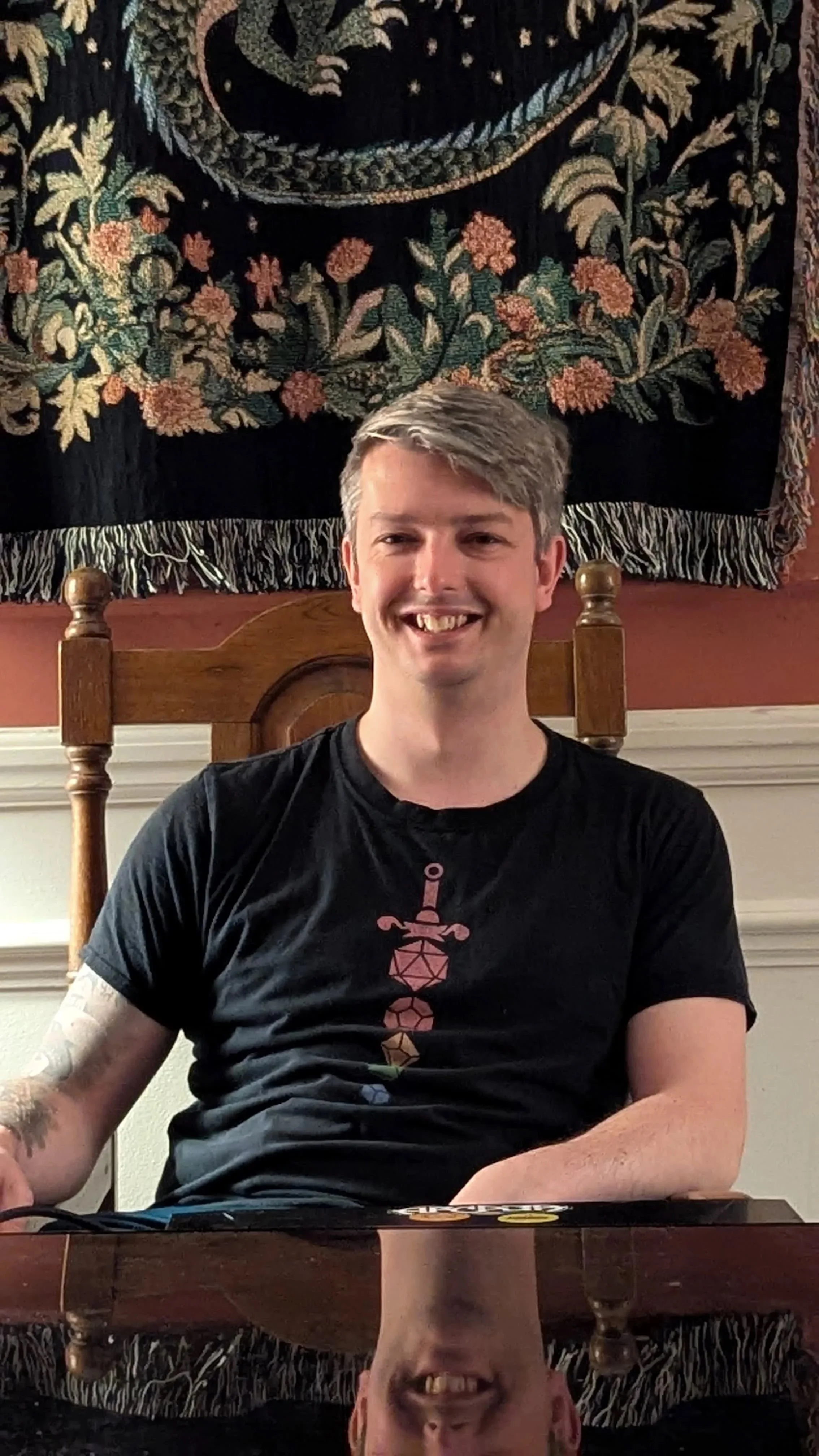 A smiling young man with short gray hair, wearing a black t-shirt with a colorful D&D sword graphic, sitting at a wooden table in a room with a tapestry of floral pattern hanging on the wall behind him.