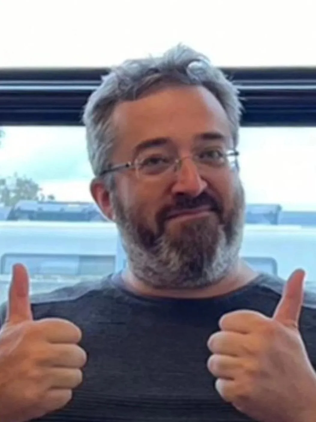 Man with gray hair and beard giving two thumbs up, sitting indoors near a window.