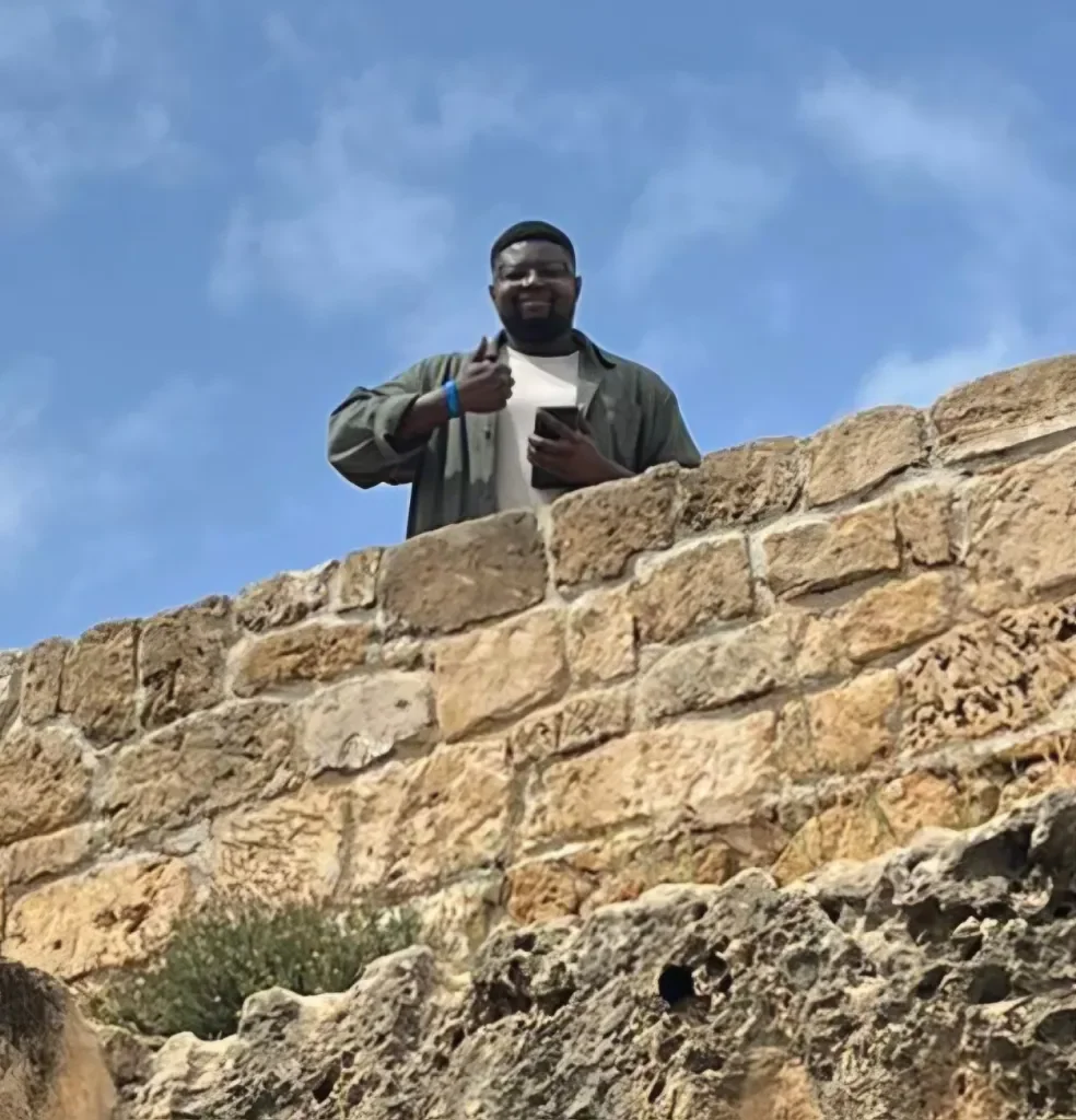 Image of a black man doing a thumbs up and smiling from far away, above an old brick wall.