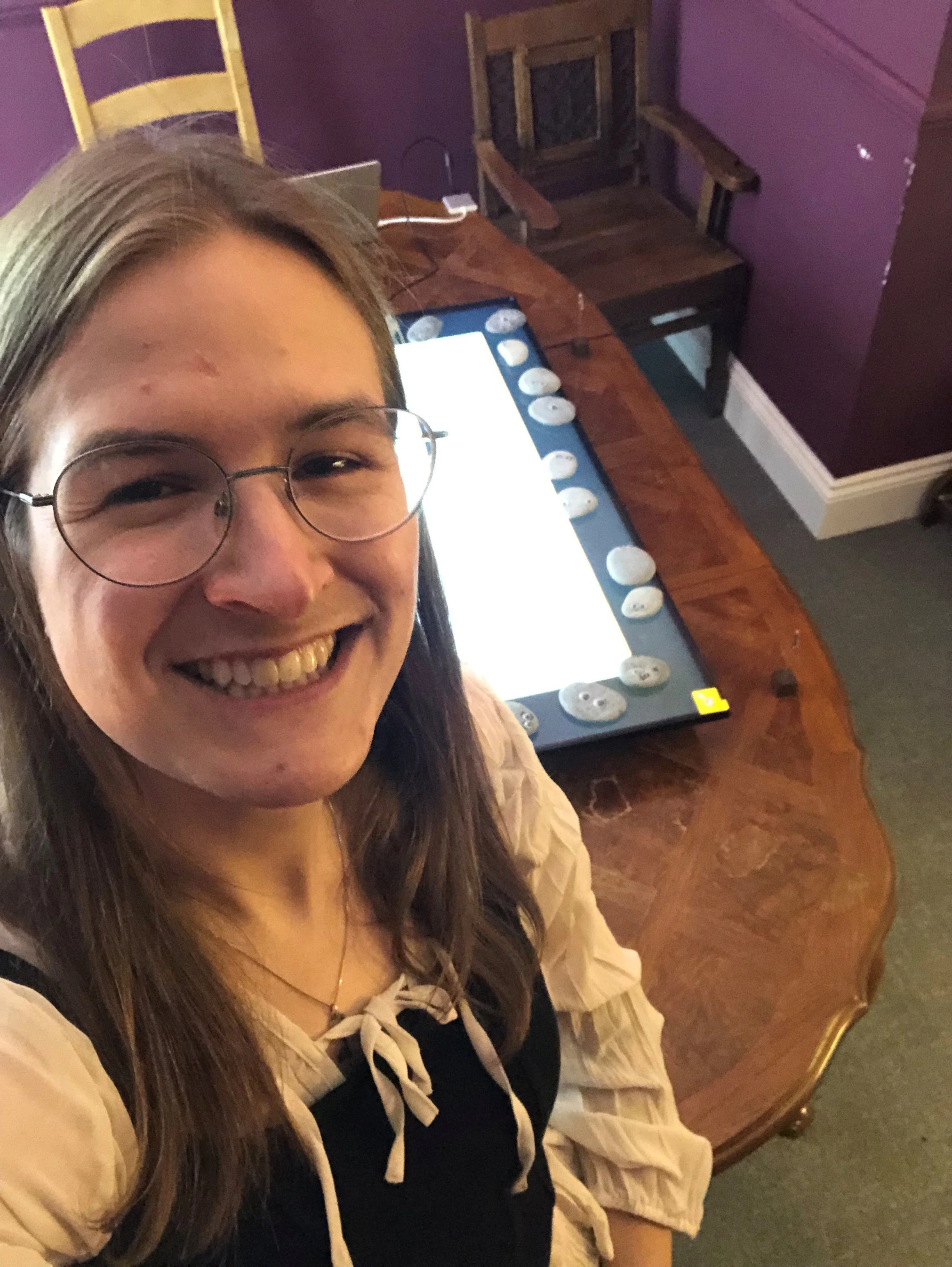 A young woman with glasses smiling in a selfie, standing in front of a large digital drawing tablet on a wooden table, with a chair and purple walls in the background.