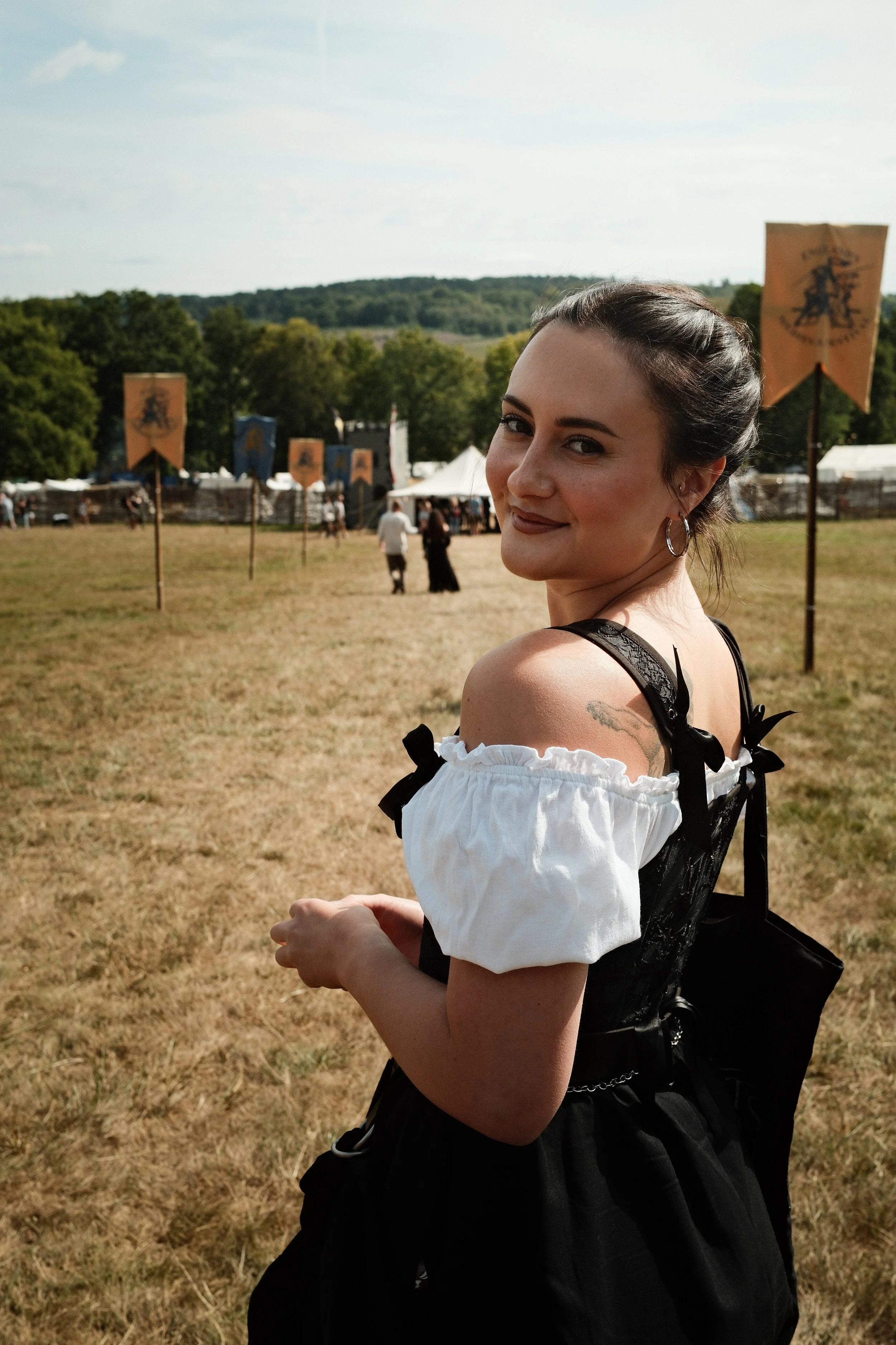 A young woman with dark hair in a bun, wearing a white off-shoulder blouse with black ribbon accents, smiling and looking over her shoulder at a medieval festival in an open field with banners and tents.