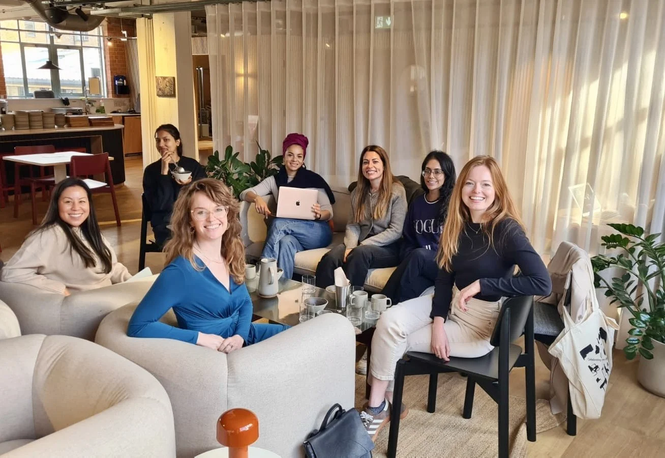 Seven women sitting and standing around a coffee table in a cozy, well-lit room, smiling and enjoying each other's company.