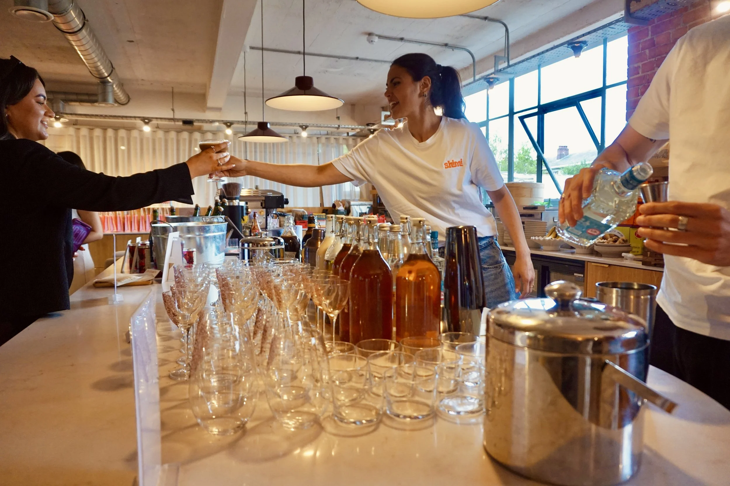 People at a bar or cafe preparing drinks, with a row of empty glasses and bottles on the counter.