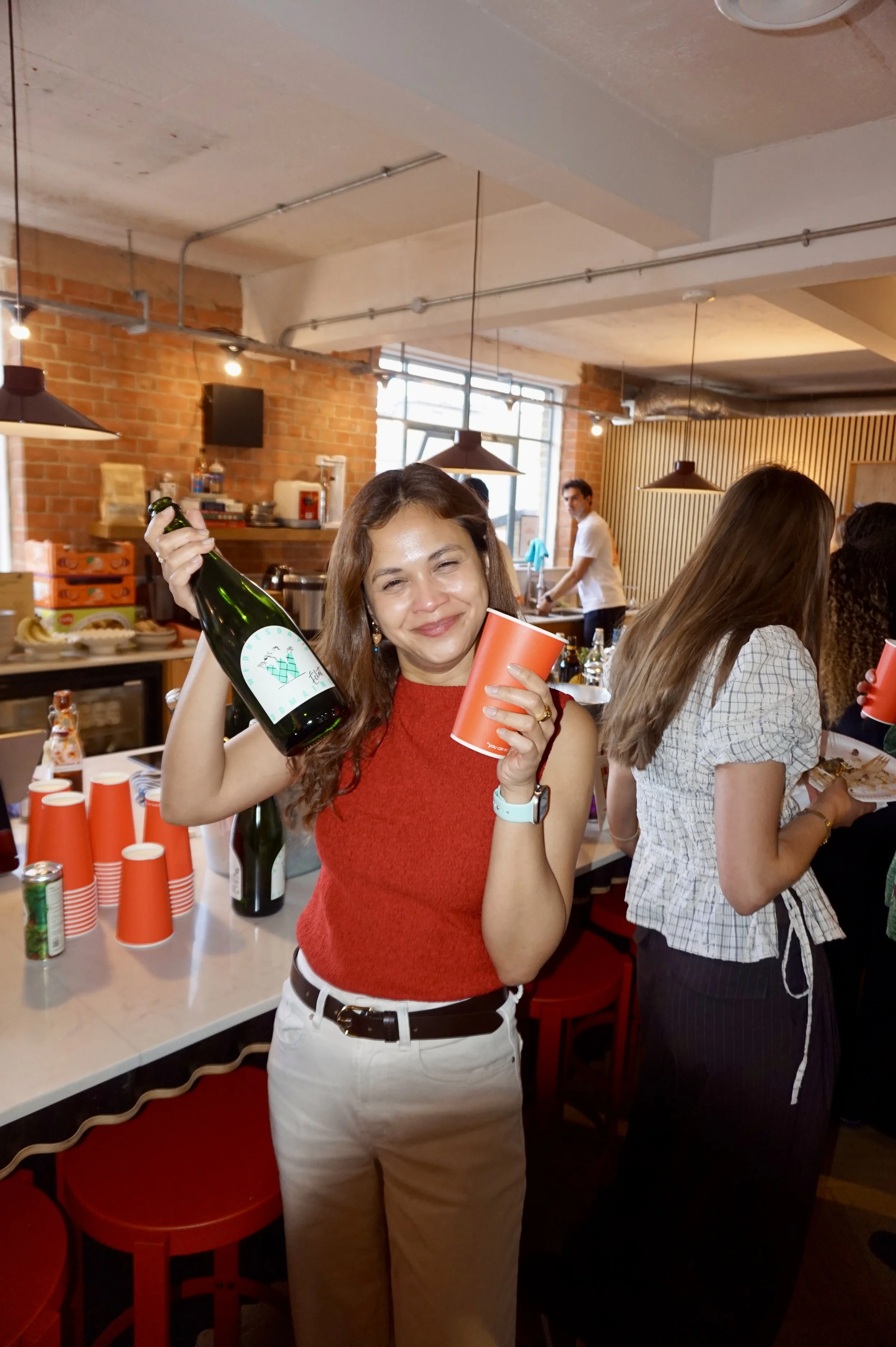 A woman with brown hair holding a wine bottle and a red cup, smiling at a celebration or gathering in a modern restaurant or bar setting with brick walls and other people in the background.