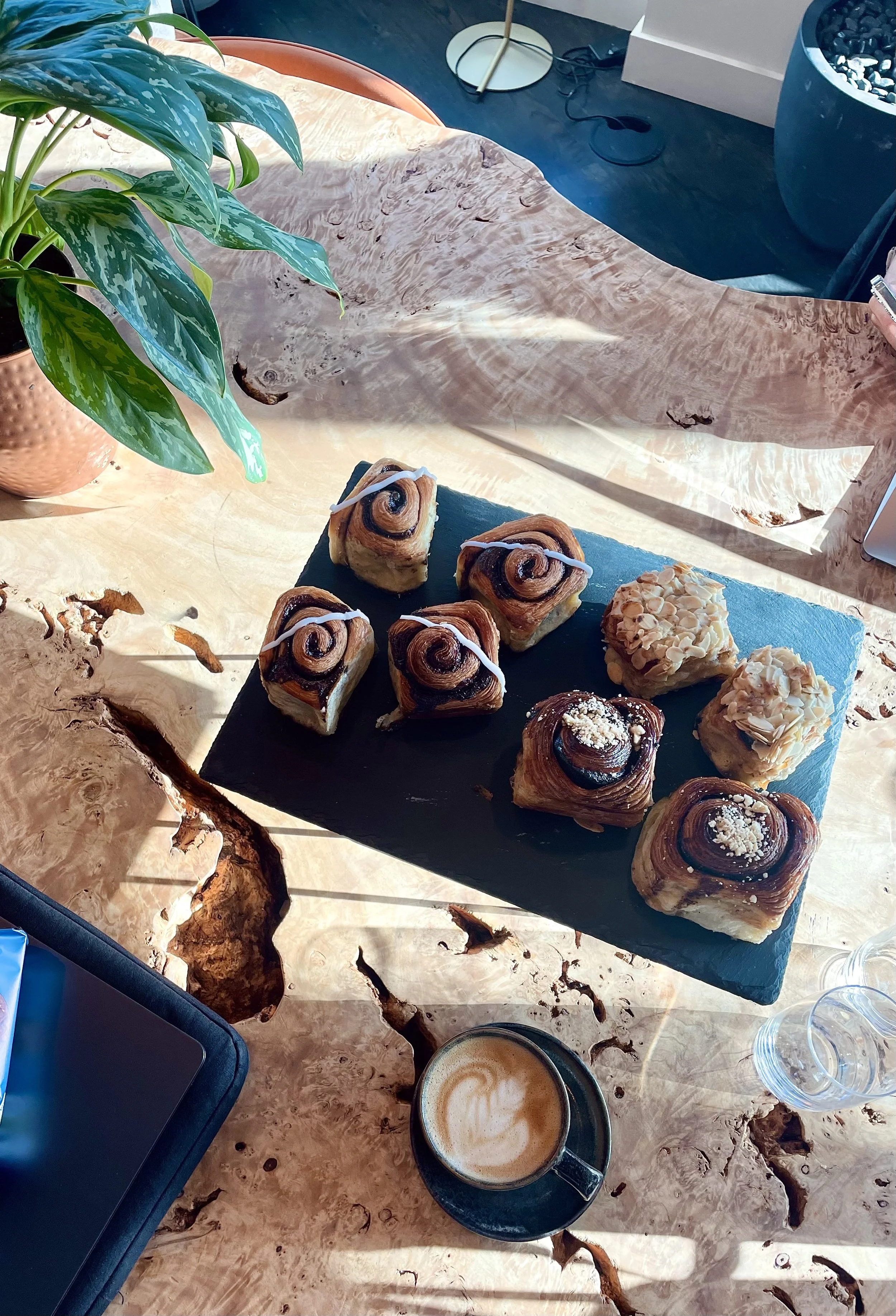 A terrazzo table with two cups of coffee with latte art, a small plate of three crumbly pastries or bars, two glasses of dark soda, and a small vase with yellow flowers. A person's lap wearing blue jeans is visible at the top of the image, with a smartphone resting on their lap.