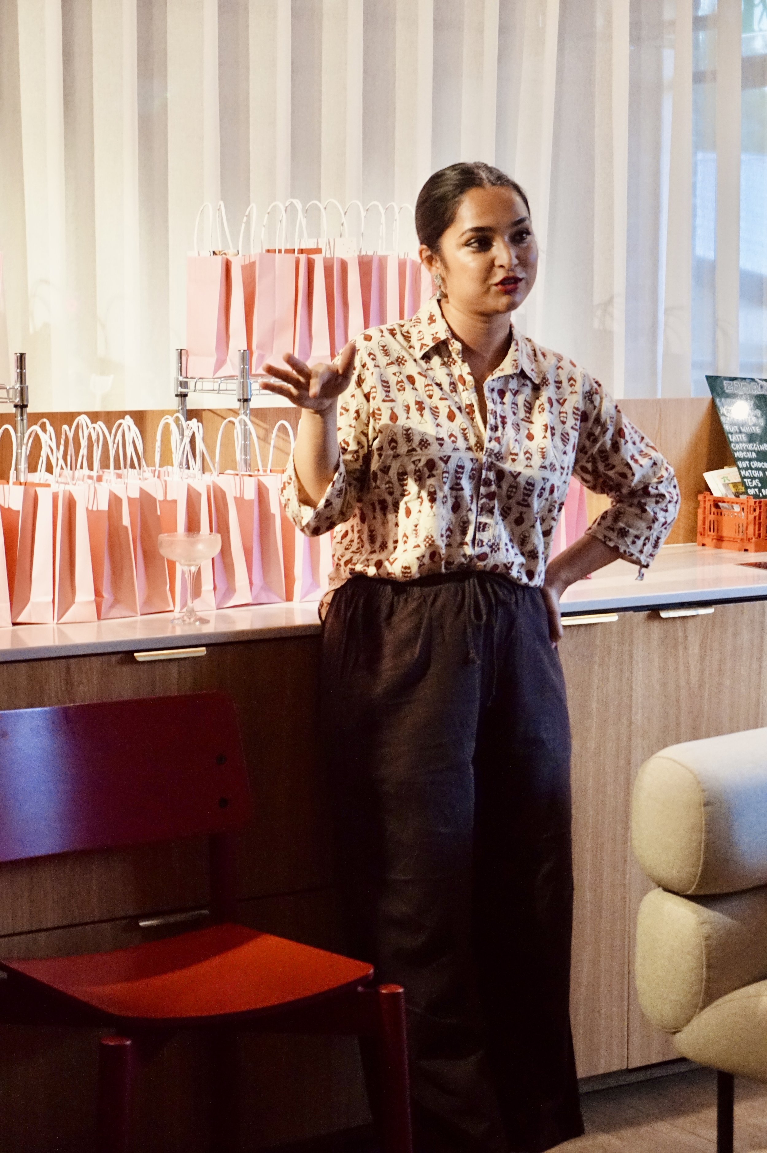 A woman with dark hair pulled back, wearing a patterned shirt and dark pants, standing inside a cafe or restaurant. Behind her are pink and white shopping bags on a counter, a menu board, and some chairs.